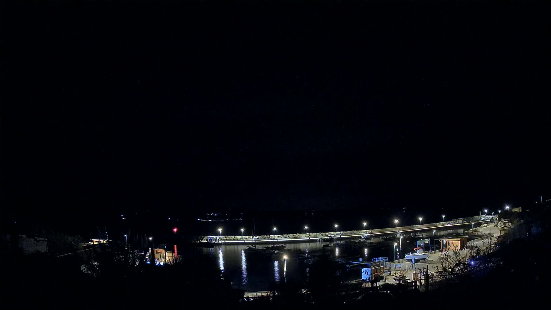 A nighttime, dimly lit harbor scene shows a curved waterfront with boats docked along a seawall, illuminated by lights.