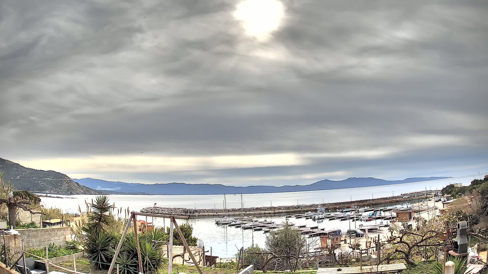 A small harbor filled with boats is seen from a hillside garden under a cloudy sky, with mountains visible across the calm water.