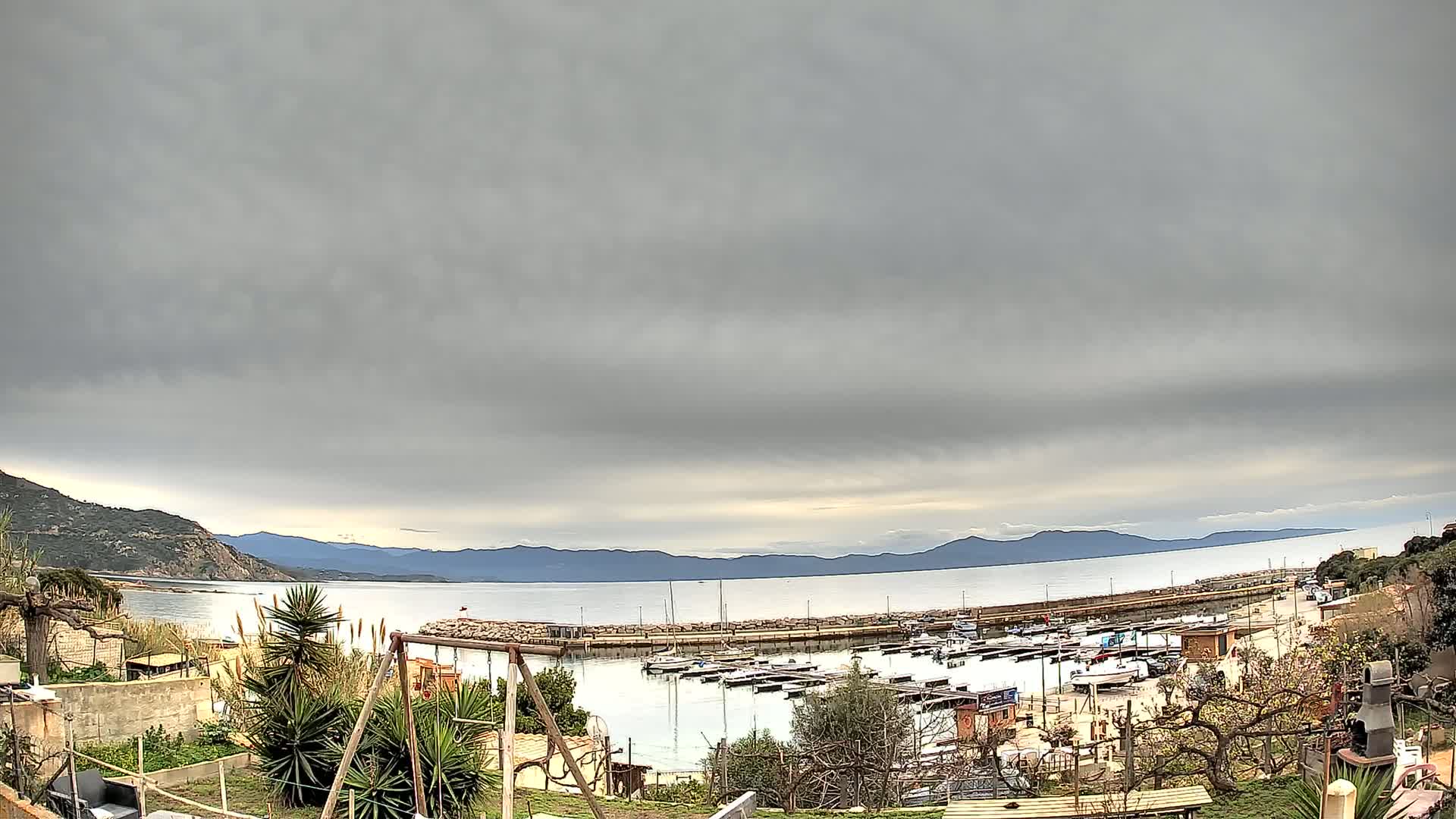 A small harbor filled with boats is seen from a garden overlooking a calm sea under an overcast sky, with mountains visible in the distance.