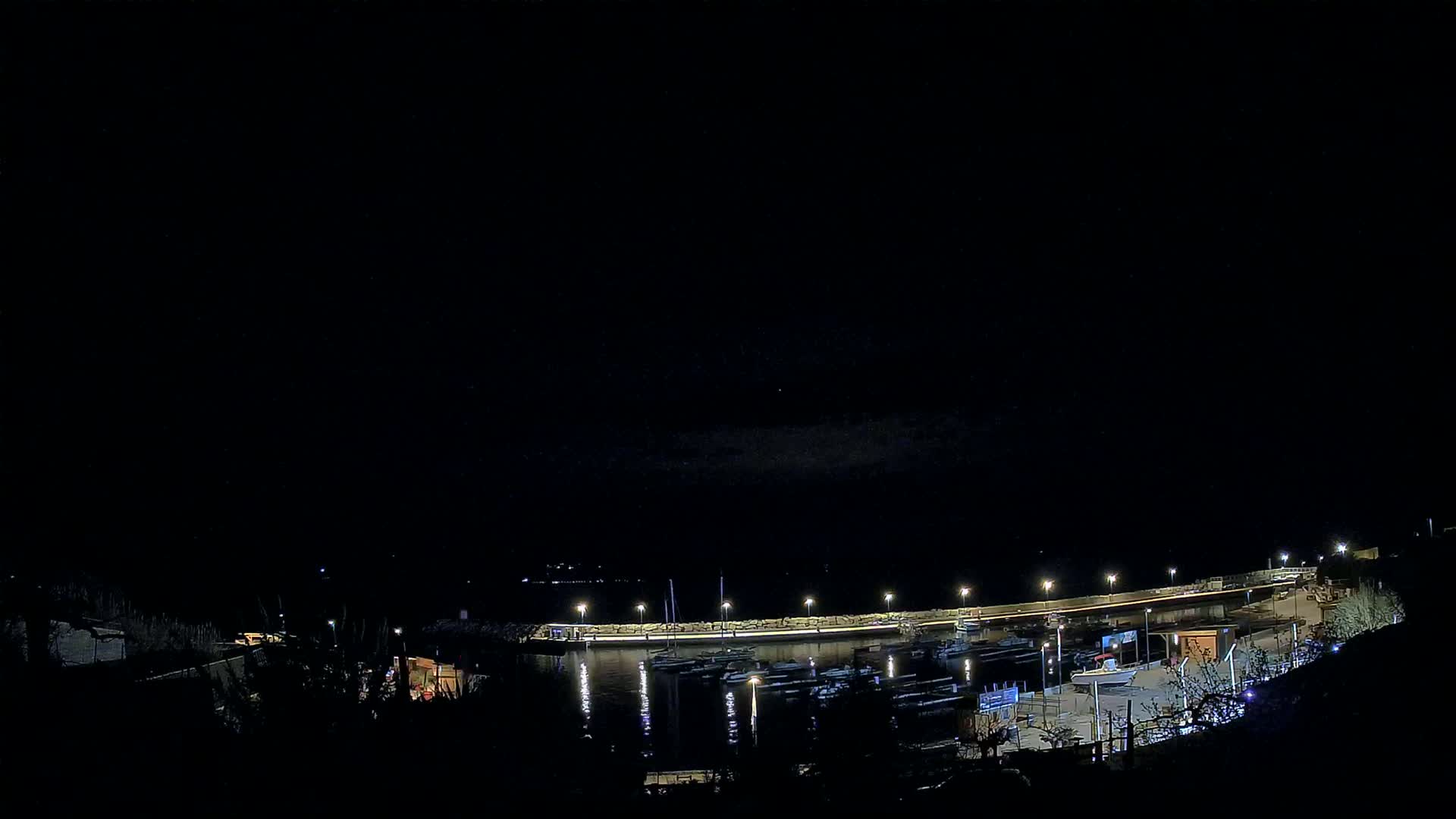 A nighttime view of a harbor filled with boats, illuminated by lights along a curved wall under a dark sky.