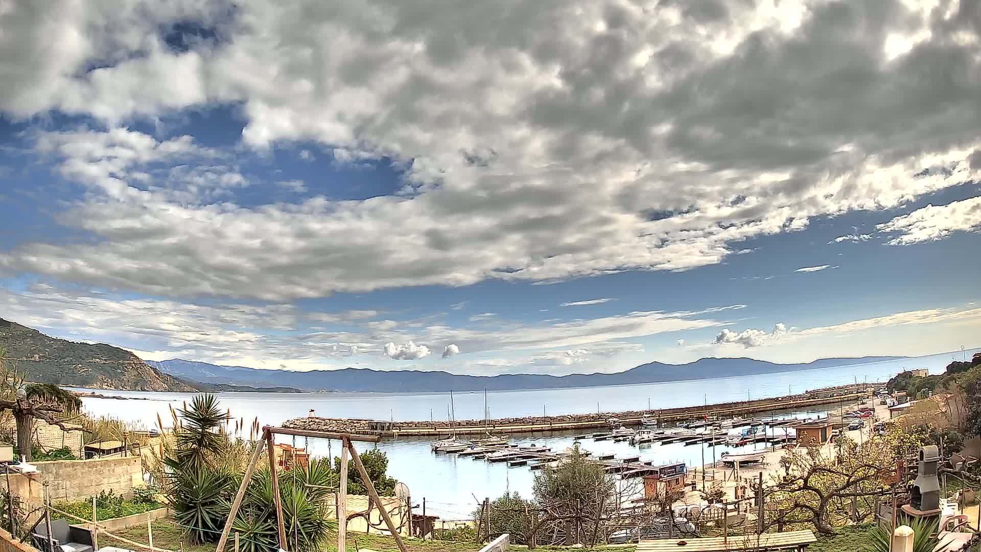 A partly cloudy sky overlooks a calm bay with a small harbor filled with boats, mountains in the distance, and a garden in the foreground.