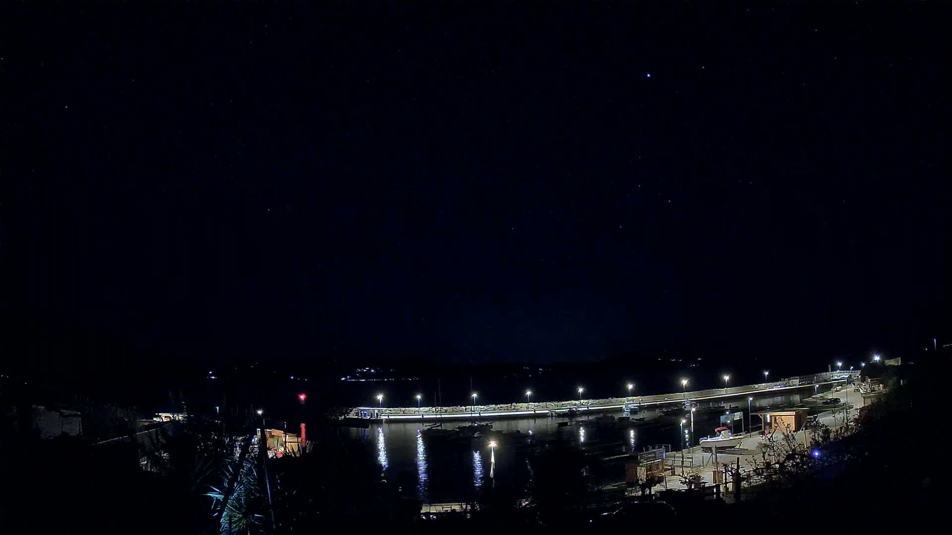 A nighttime view of a harbor with boats docked along a lit seawall under a clear, starry sky.