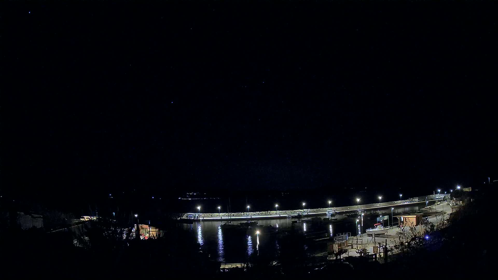 A nighttime view of a harbor with boats docked, illuminated by lights under a clear, starlit sky.