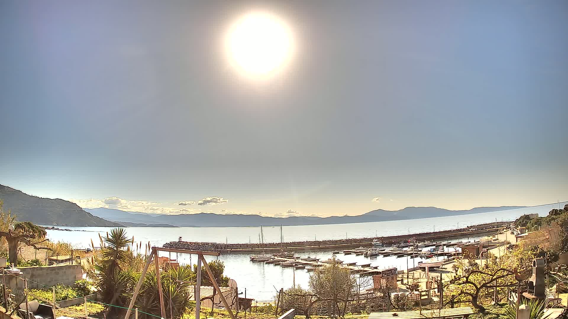 A sunny day reveals a harbor filled with boats, nestled between a tranquil sea and distant mountains.