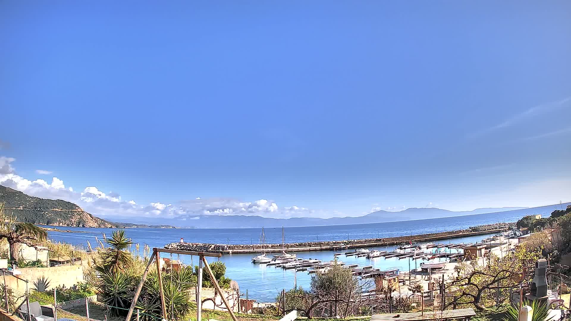 A mostly sunny day reveals a small harbor filled with boats, nestled between a hillside and calm blue ocean under a clear sky.
