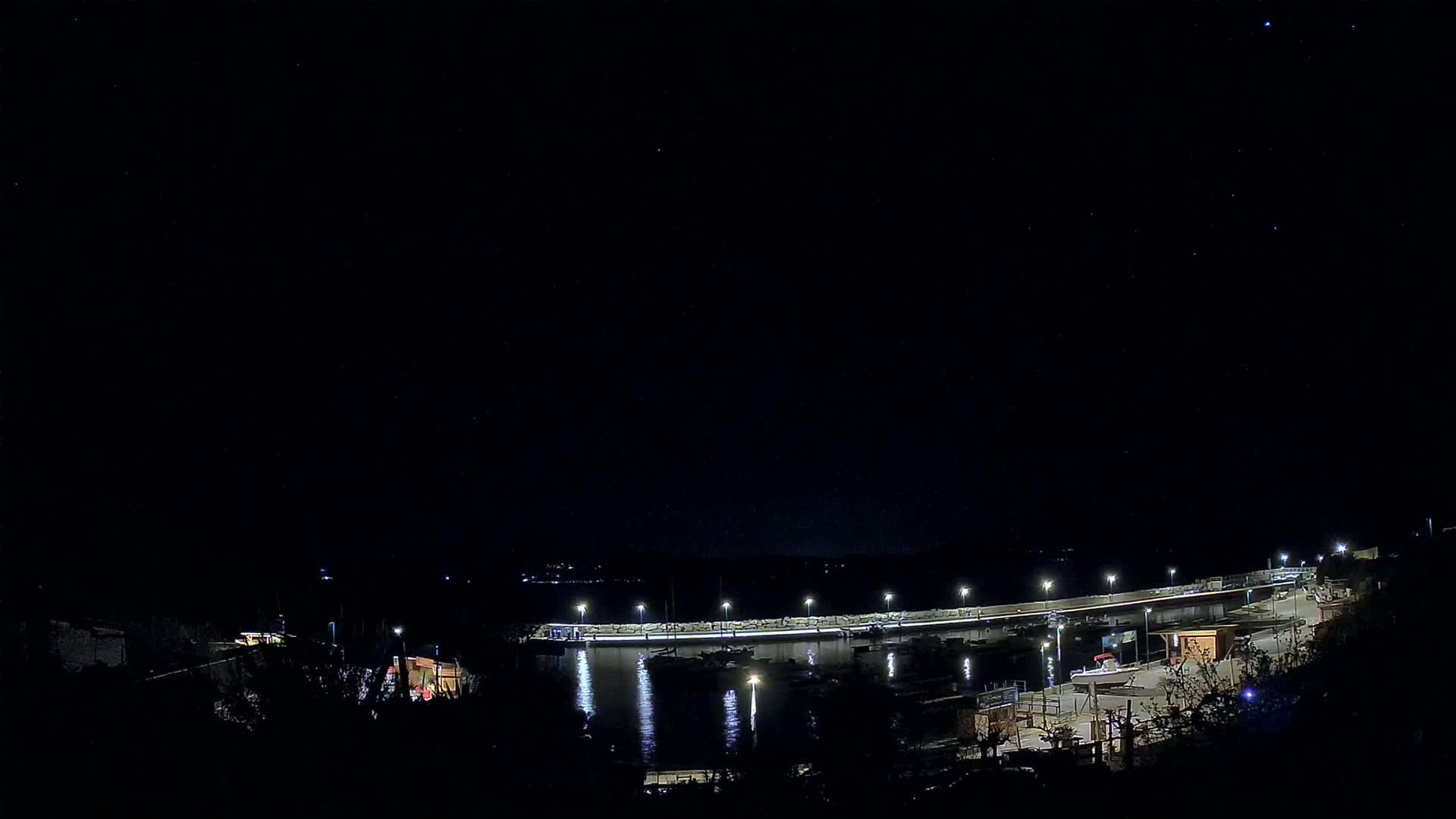 A nighttime view of a harbor with boats docked, illuminated by lights against a clear, starry sky.
