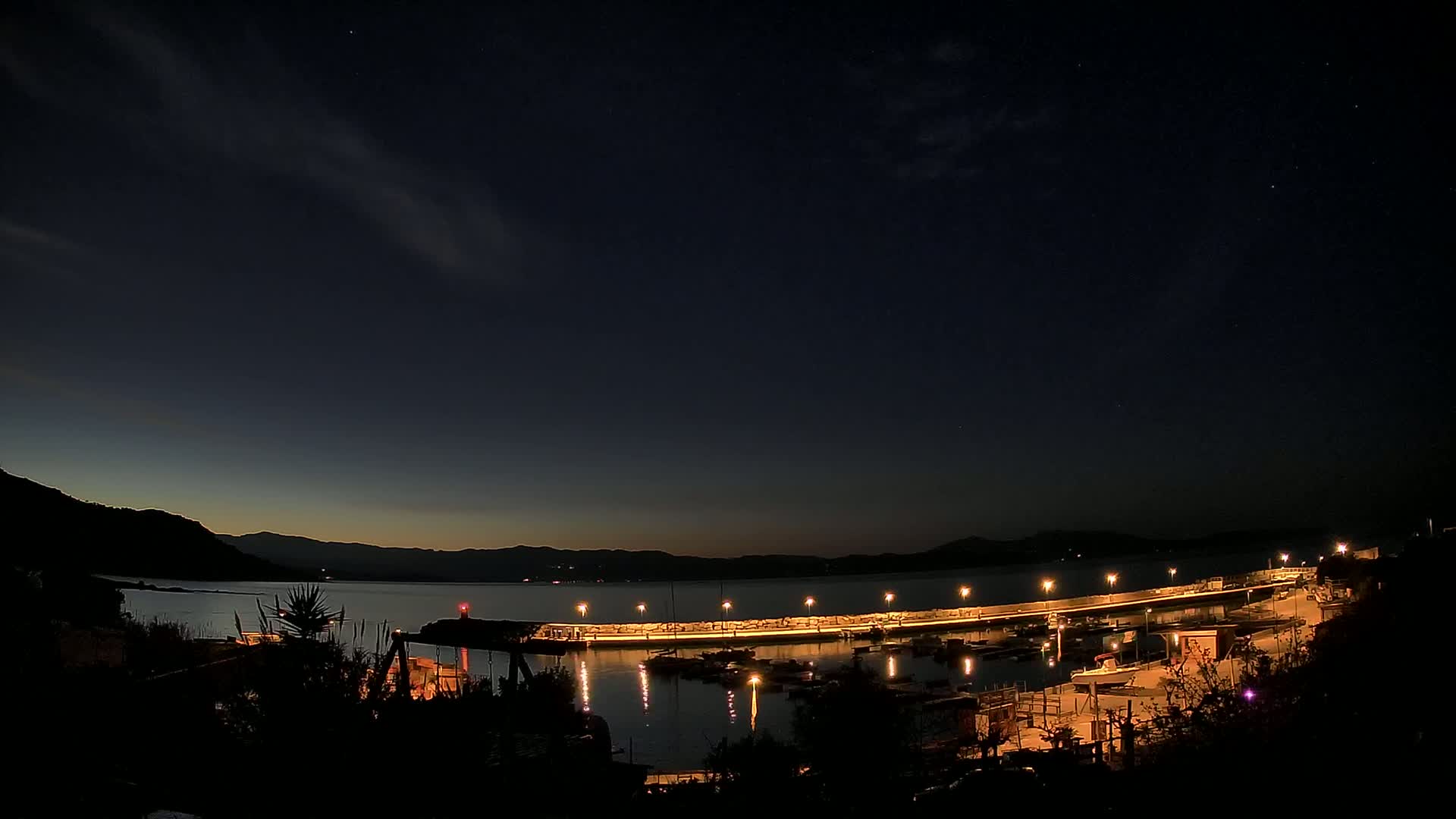 A nighttime view of a harbor with boats and lit docks, under a dark sky with some clouds and distant mountains.