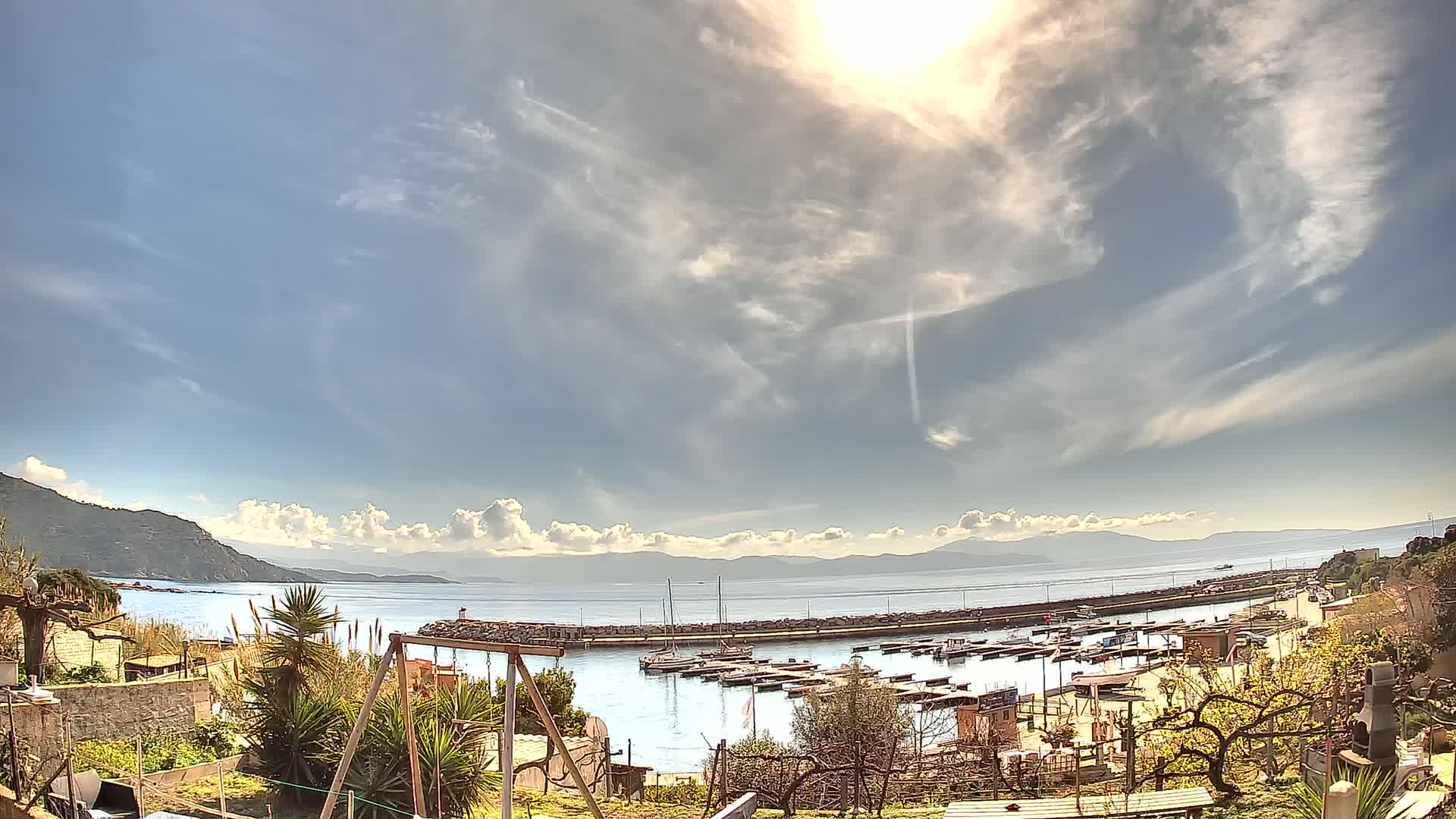A sunny day with partly cloudy skies overlooks a calm harbor filled with boats, nestled between a hillside and distant mountains.