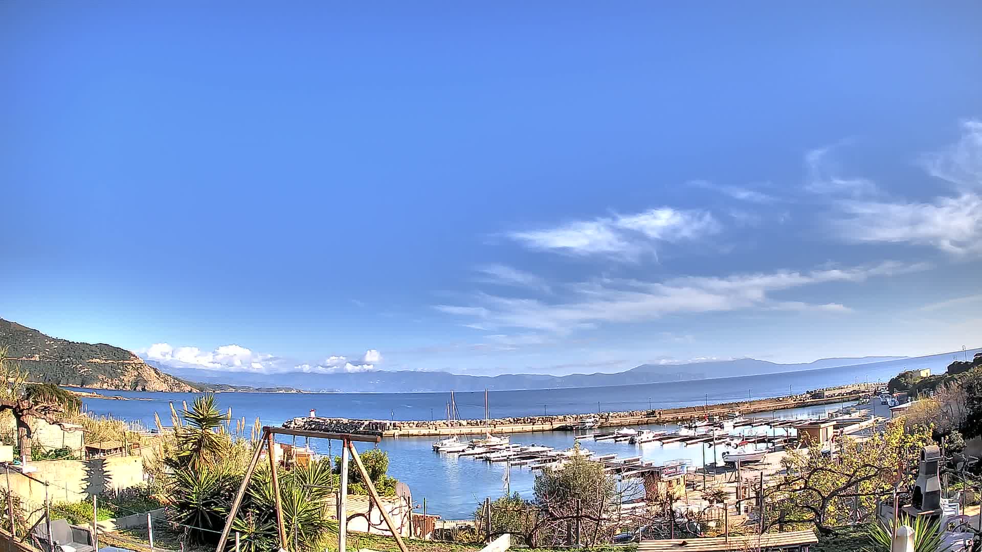 A small harbor filled with boats is nestled between a rocky hillside and calm blue water under a mostly sunny sky with scattered clouds.