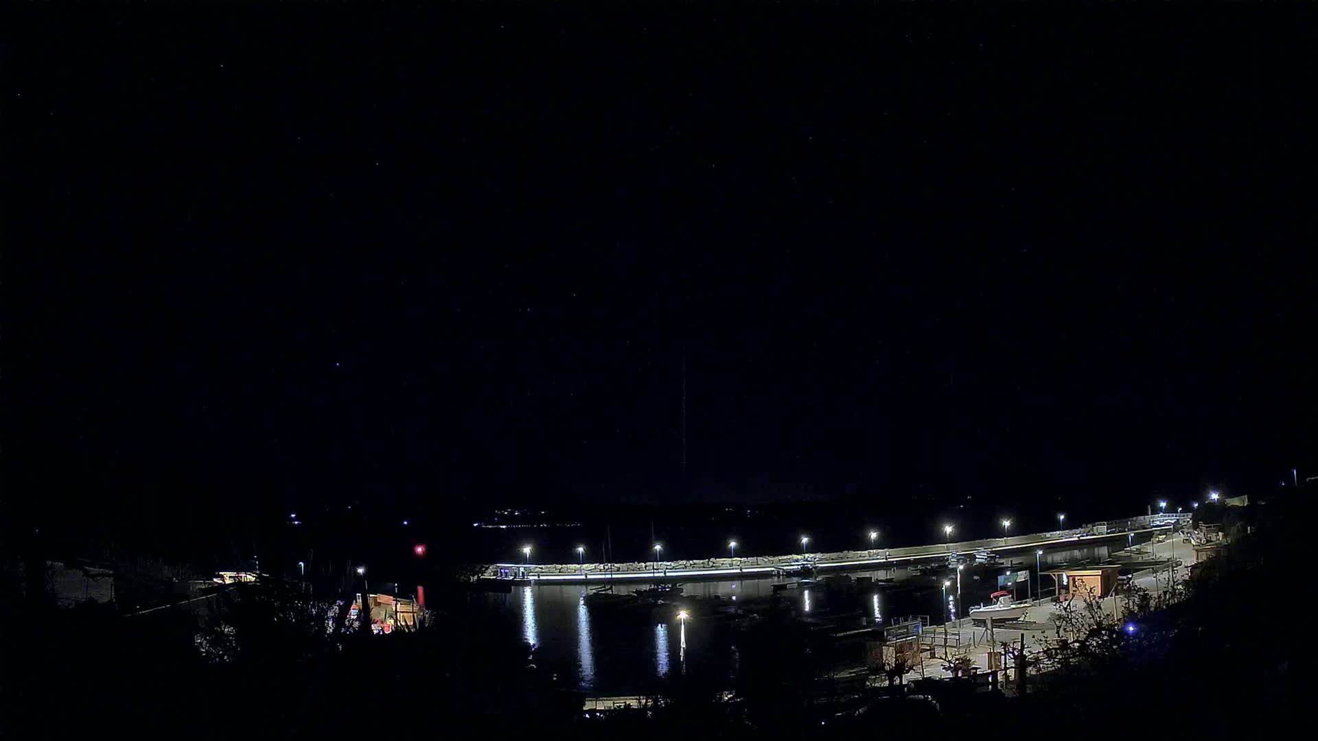A nighttime view of a harbor with boats moored along a lit seawall under a clear, starry sky.