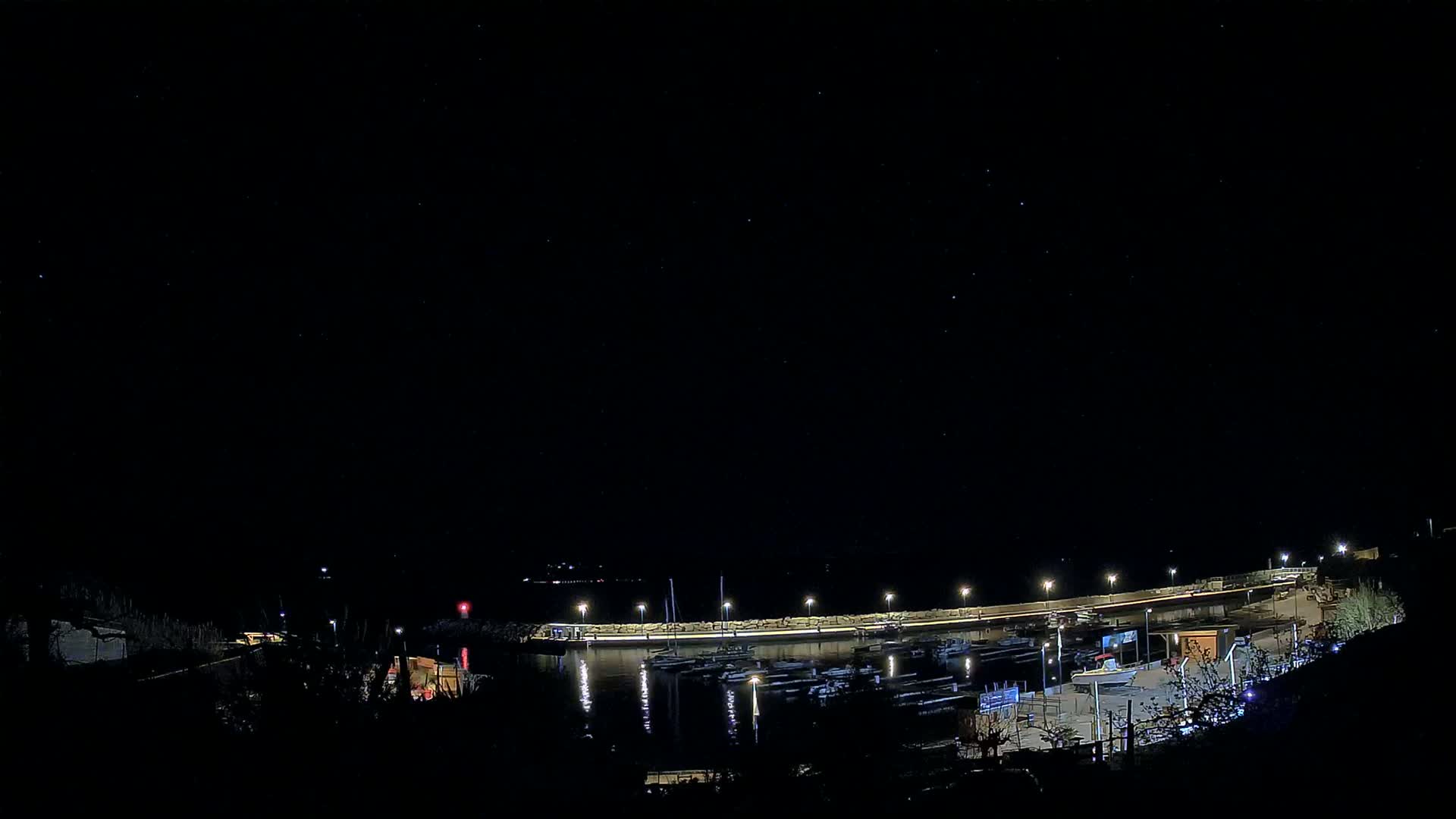A nighttime view of a marina with numerous boats docked, illuminated by lights under a clear, starry sky.