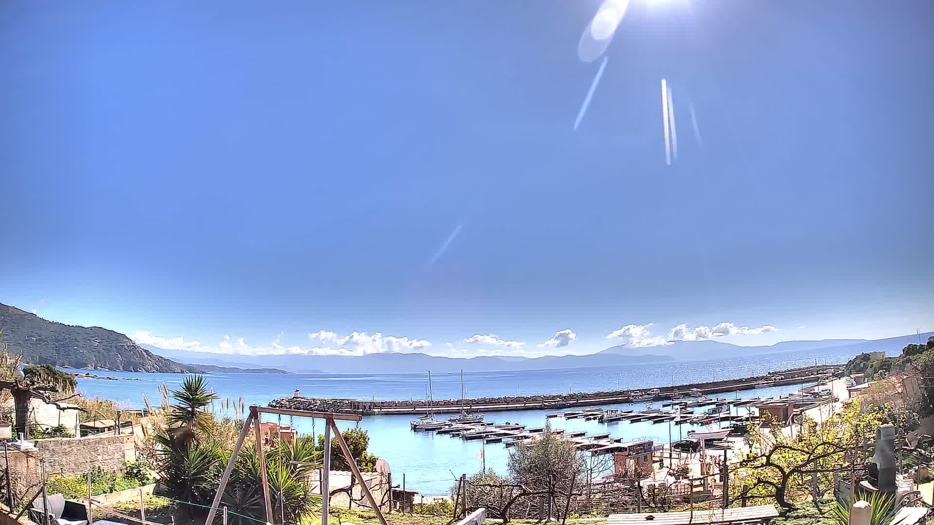 A sunny day reveals a small harbor filled with boats, nestled between a rocky coastline and a calm sea under a mostly clear blue sky.