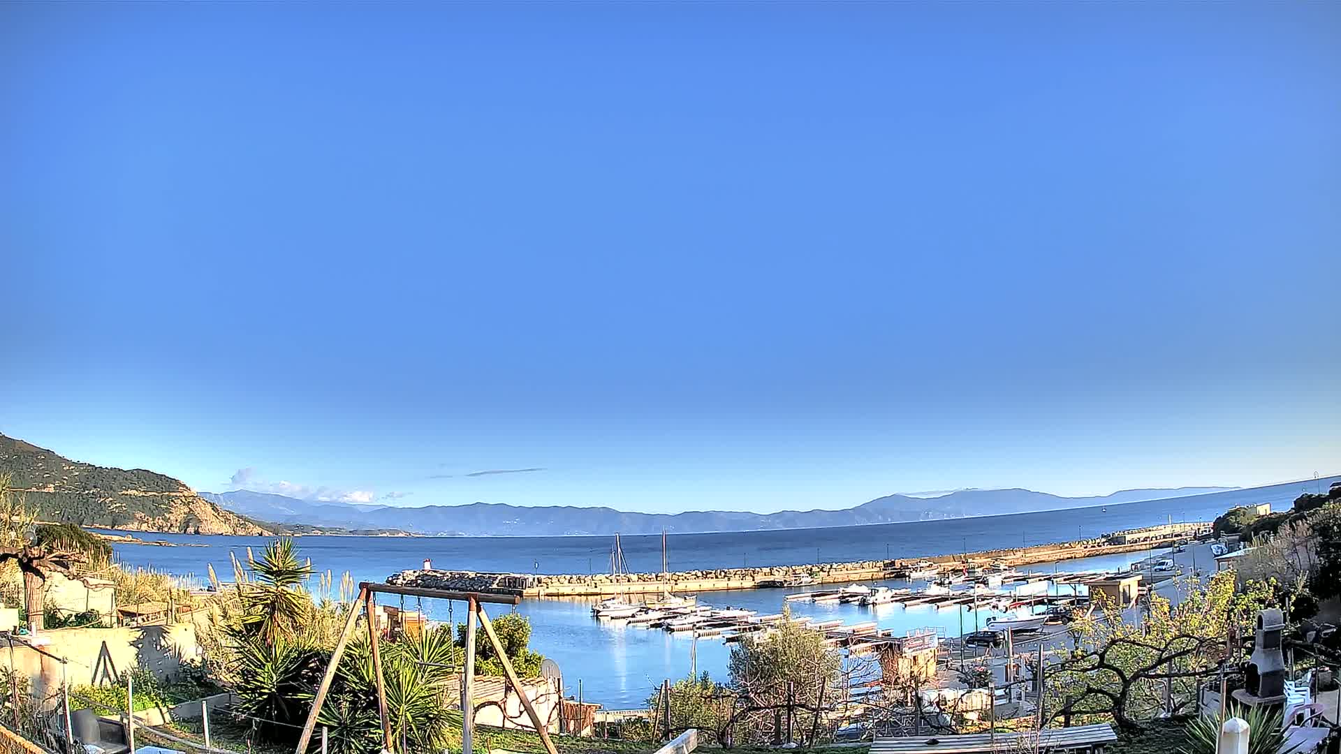A clear blue sky overlooks a calm harbor filled with boats, nestled between a rocky hillside and distant mountains.