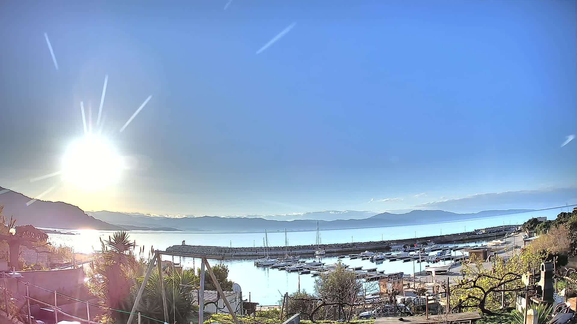 A sunny day reveals a harbor filled with boats, nestled between a calm sea and distant mountains.
