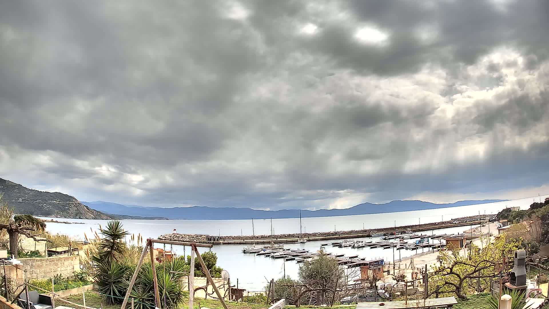 A small harbor filled with boats is nestled between a hilly coastline and a calm sea under a heavily clouded sky.