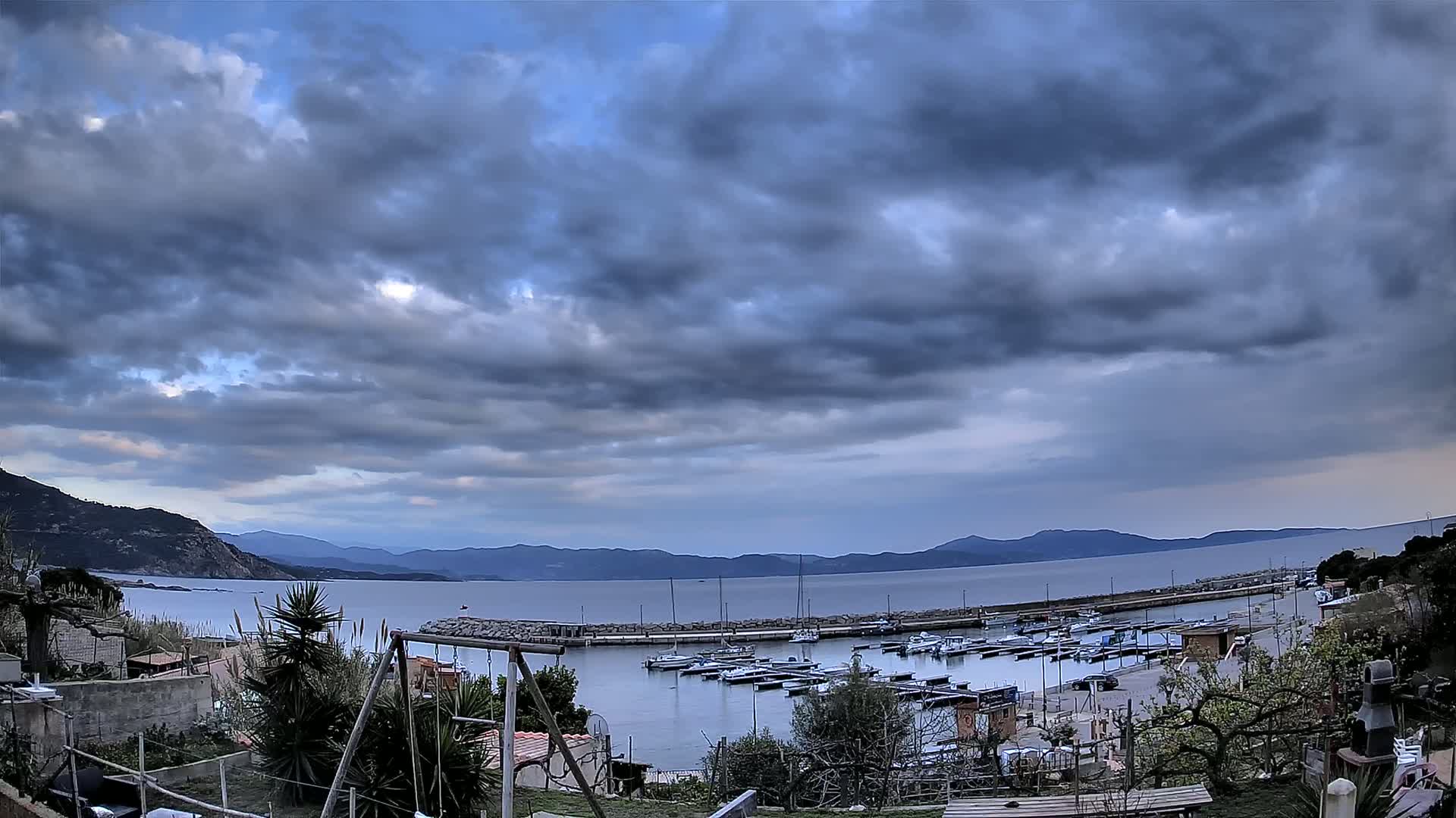 A small marina filled with boats sits under a cloudy sky, with mountains in the distance.