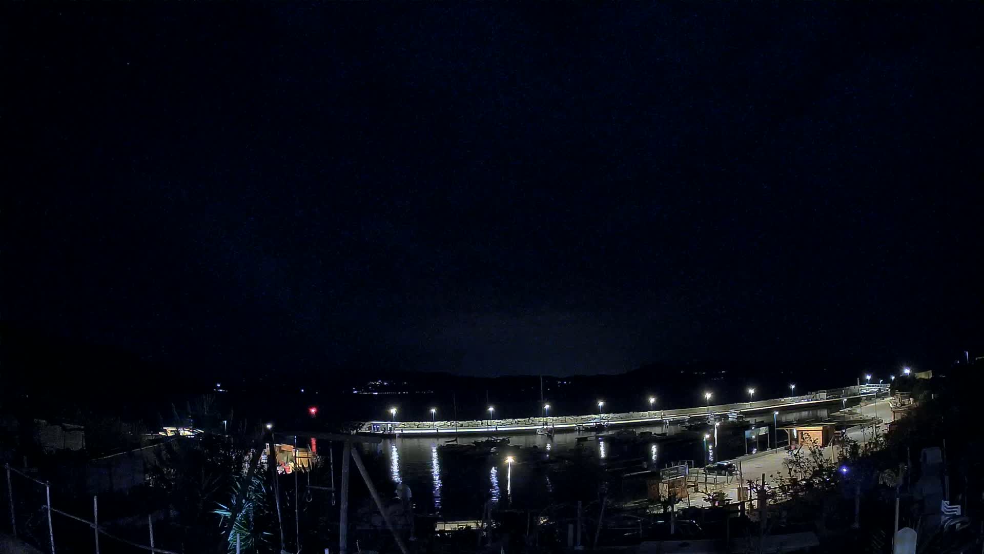 A nighttime view of a harbor with boats moored alongside a lit seawall under a clear, dark sky.