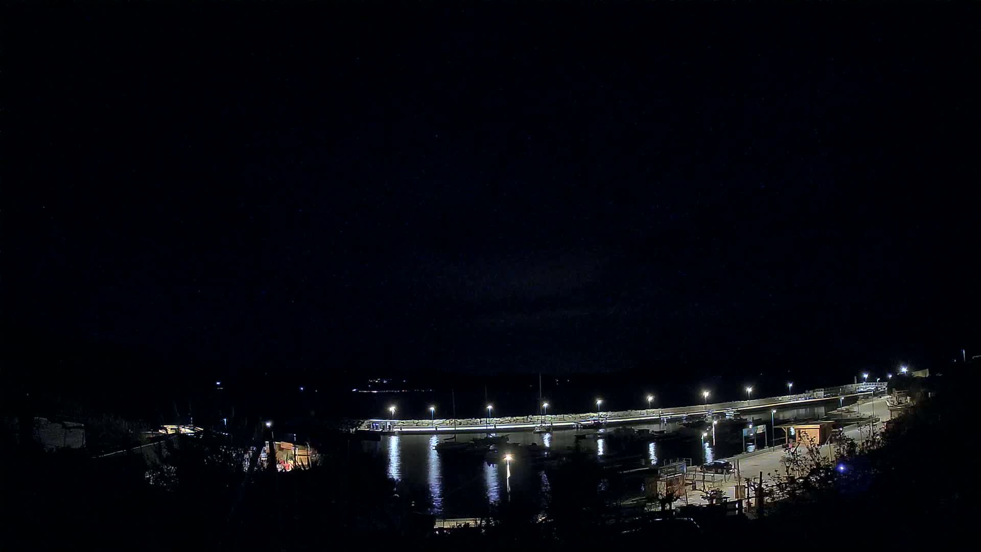 A nighttime view of a harbor with boats docked along a lit seawall under a clear, dark sky.