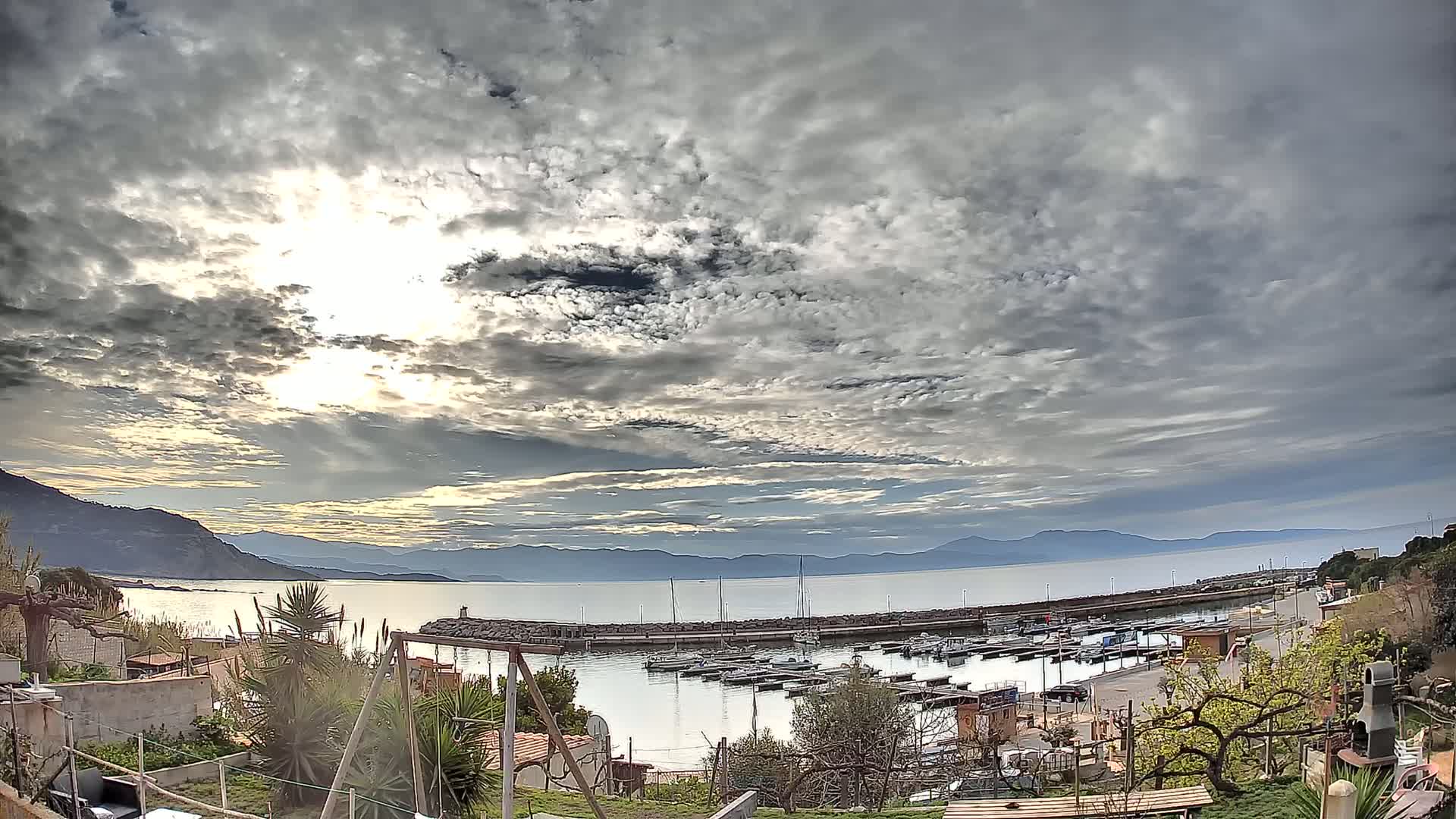 A mostly cloudy sky with bright sunshine overhead overlooks a calm bay containing a small marina, backed by mountains and a residential area with lush vegetation.