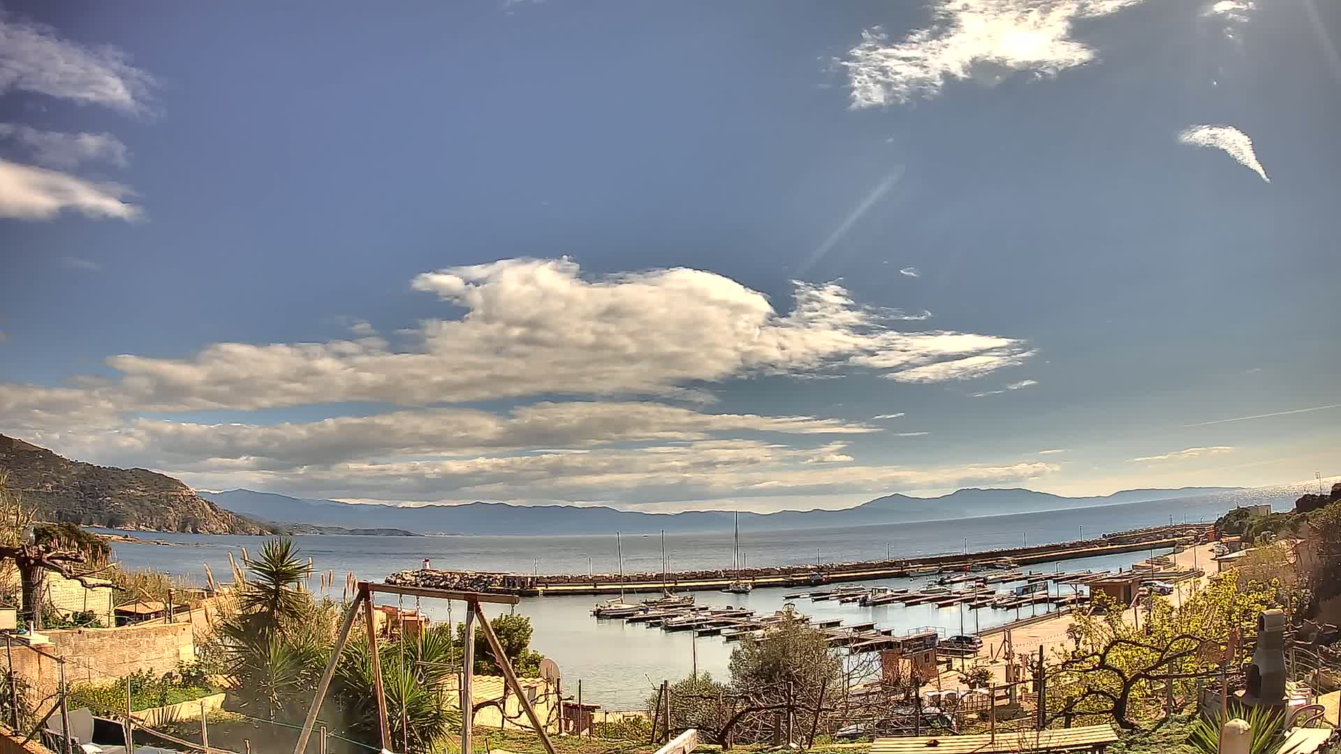 A small harbor filled with boats is nestled between a hillside and a calm sea under a partly cloudy sky.