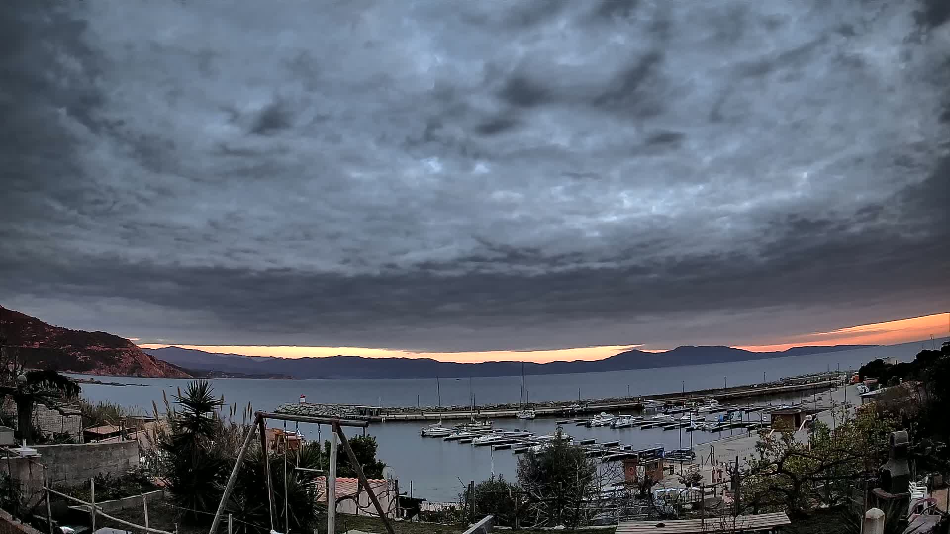 A small marina filled with boats is nestled between hills under a dark, cloudy sky with a hint of orange on the horizon.