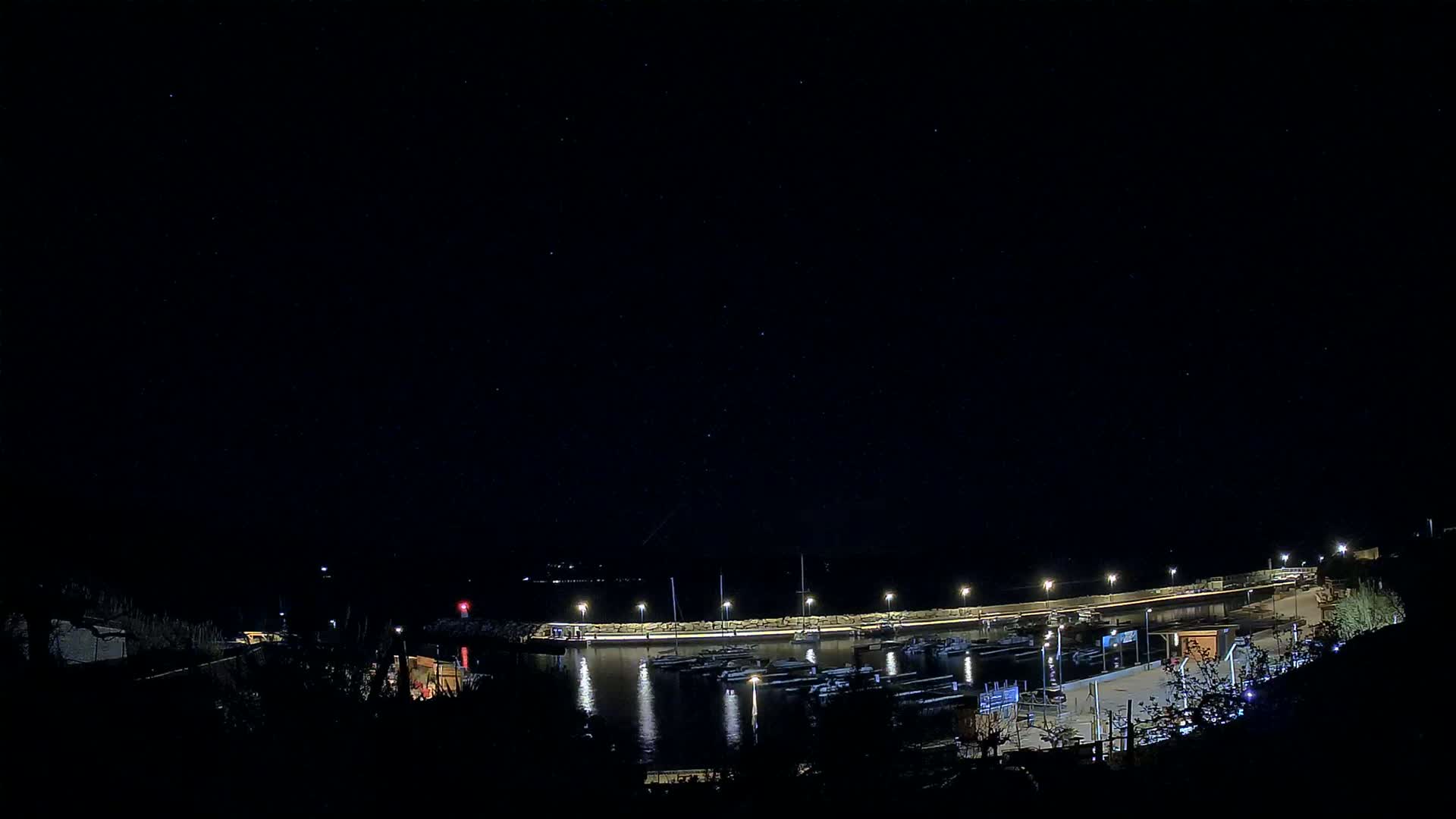 A nighttime view of a harbor filled with boats, illuminated by lights along a seawall under a clear, starry sky.