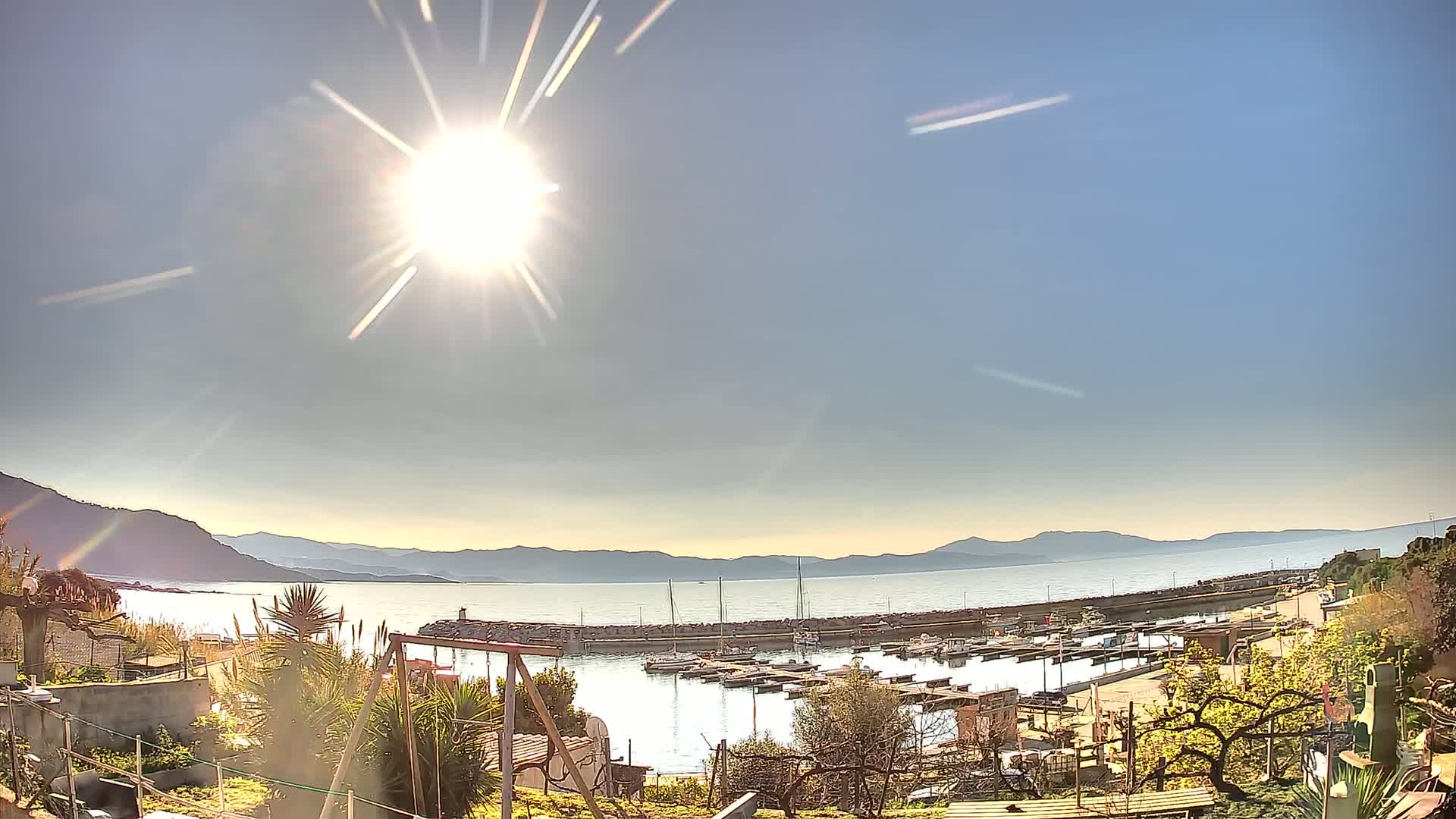 A sunny day reveals a harbor filled with boats, nestled between a hillside and a calm body of water stretching towards distant mountains.