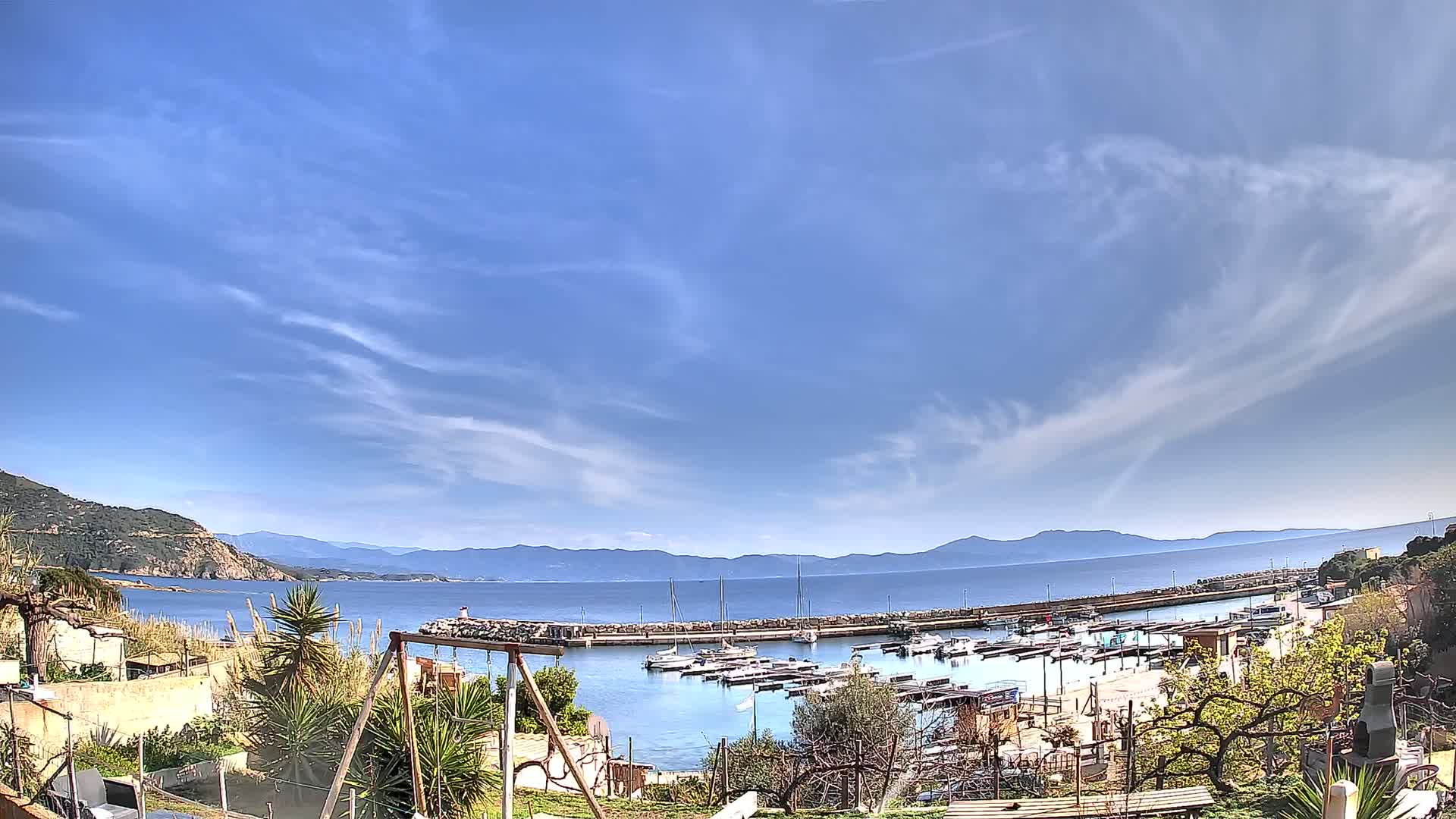 A small harbor filled with boats sits below a clear blue sky with wispy clouds, backed by a calm sea and distant mountains.