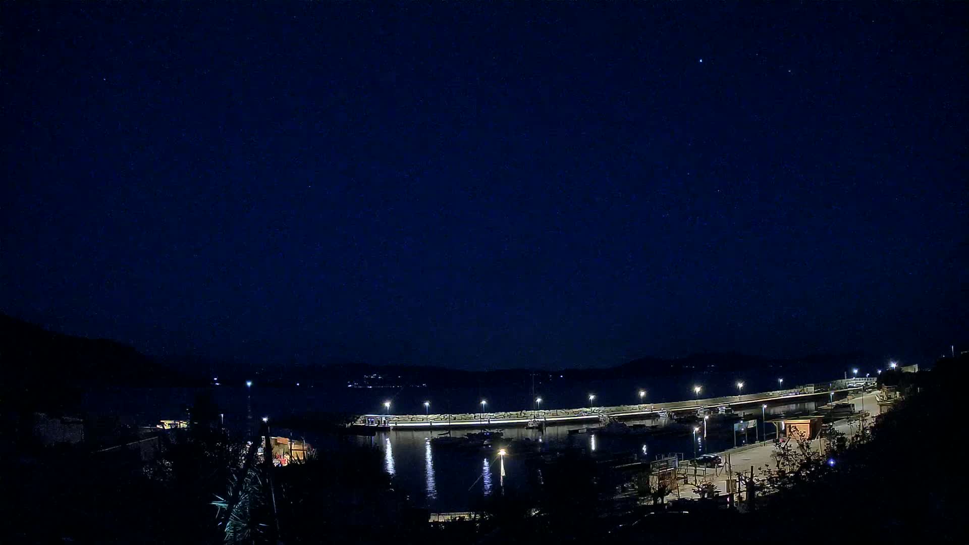 A nighttime view of a harbor with boats and a long pier illuminated under a clear, starry sky.
