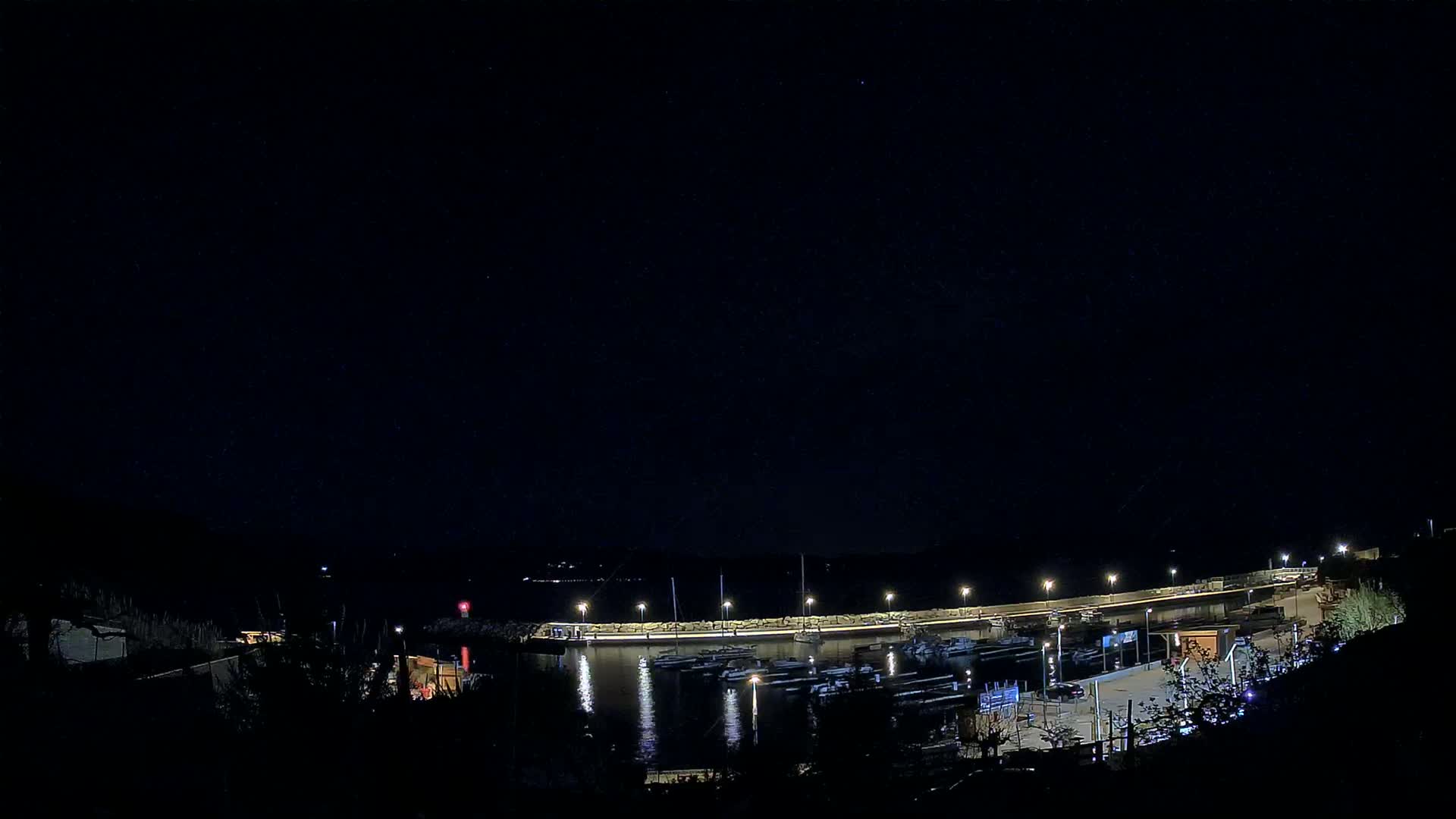 A nighttime view of a harbor filled with boats, illuminated by lights under a clear, dark sky.
