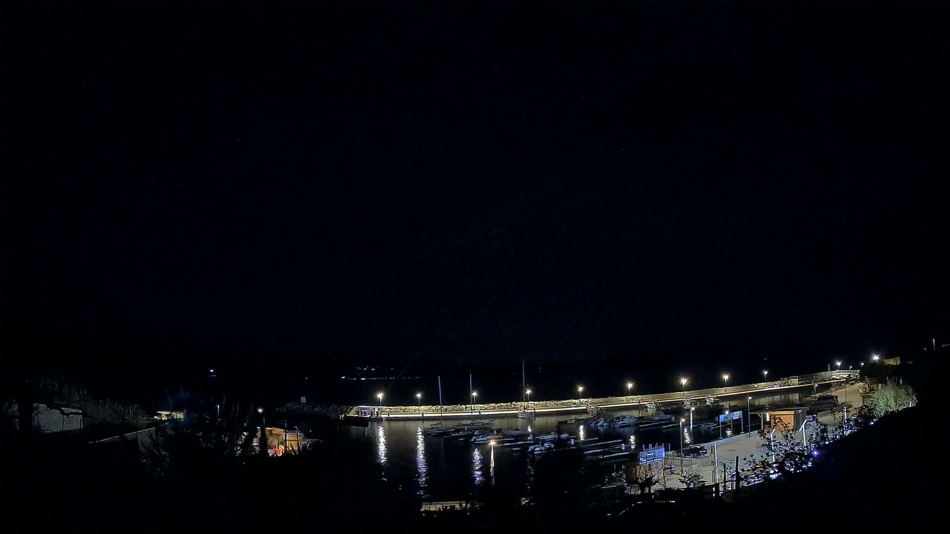 A nighttime view of a harbor filled with boats, illuminated by lights along a curved wall under a clear, dark sky.