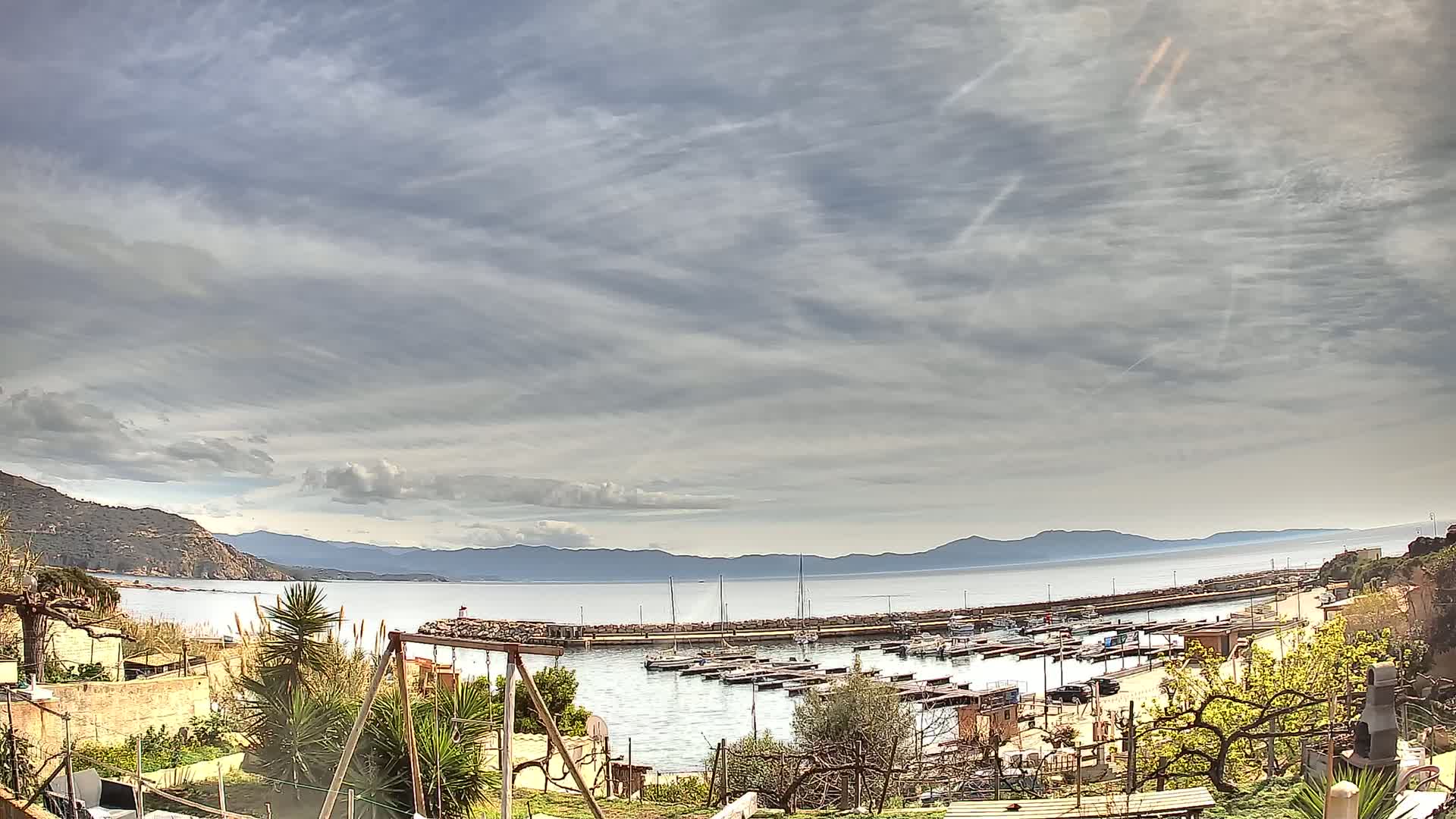 A calm bay with numerous boats in a small harbor is seen from a hillside under a mostly cloudy sky.