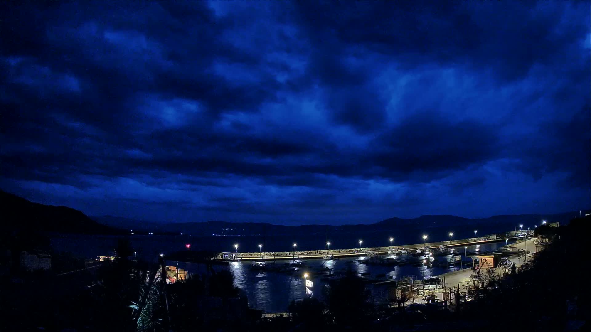 A nighttime harbor scene under a dark blue, cloudy sky shows a breakwater with docked boats and illuminated pathways.