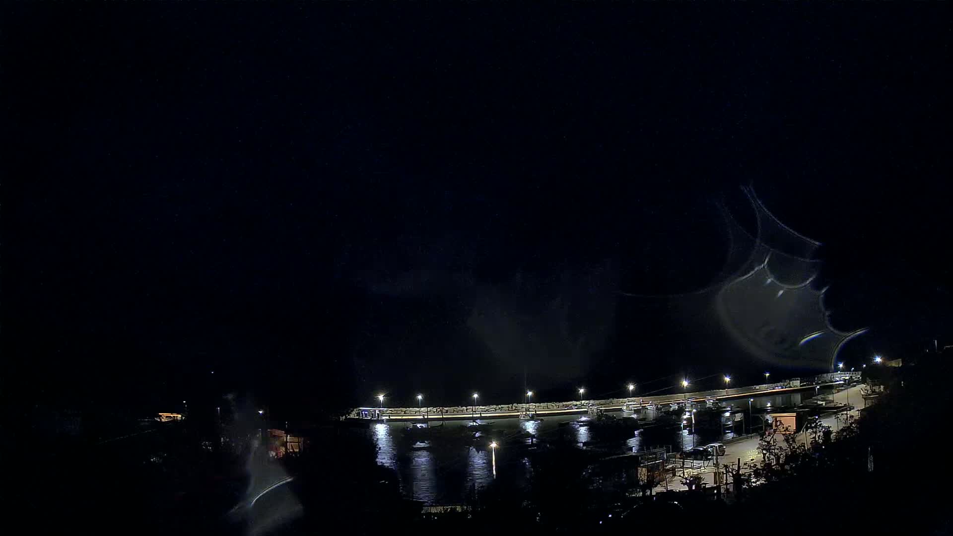 A nighttime, low-angle view of a harbor with boats, illuminated by lights, under a dark, possibly cloudy sky.