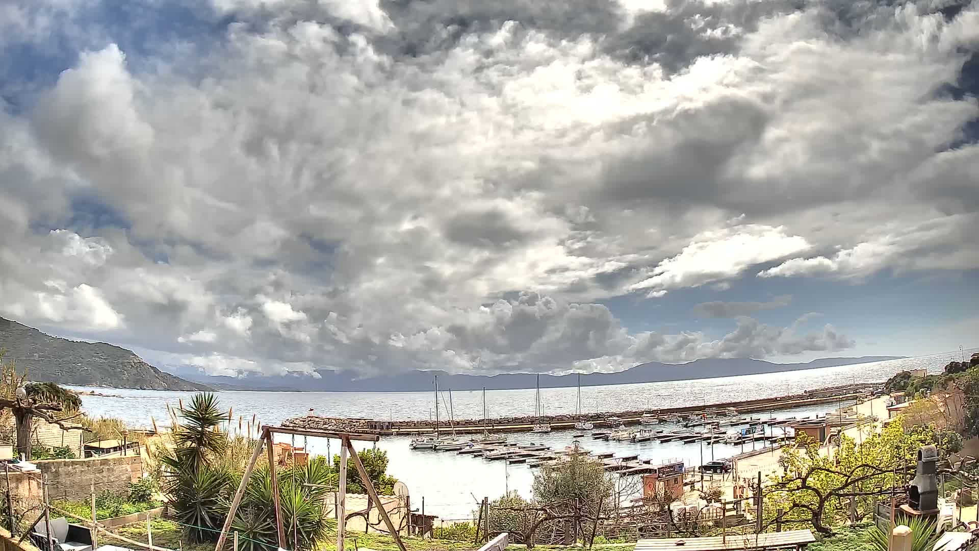 A partly cloudy day shows a small harbor filled with boats, nestled between a rocky shoreline and a calm sea with distant mountains.