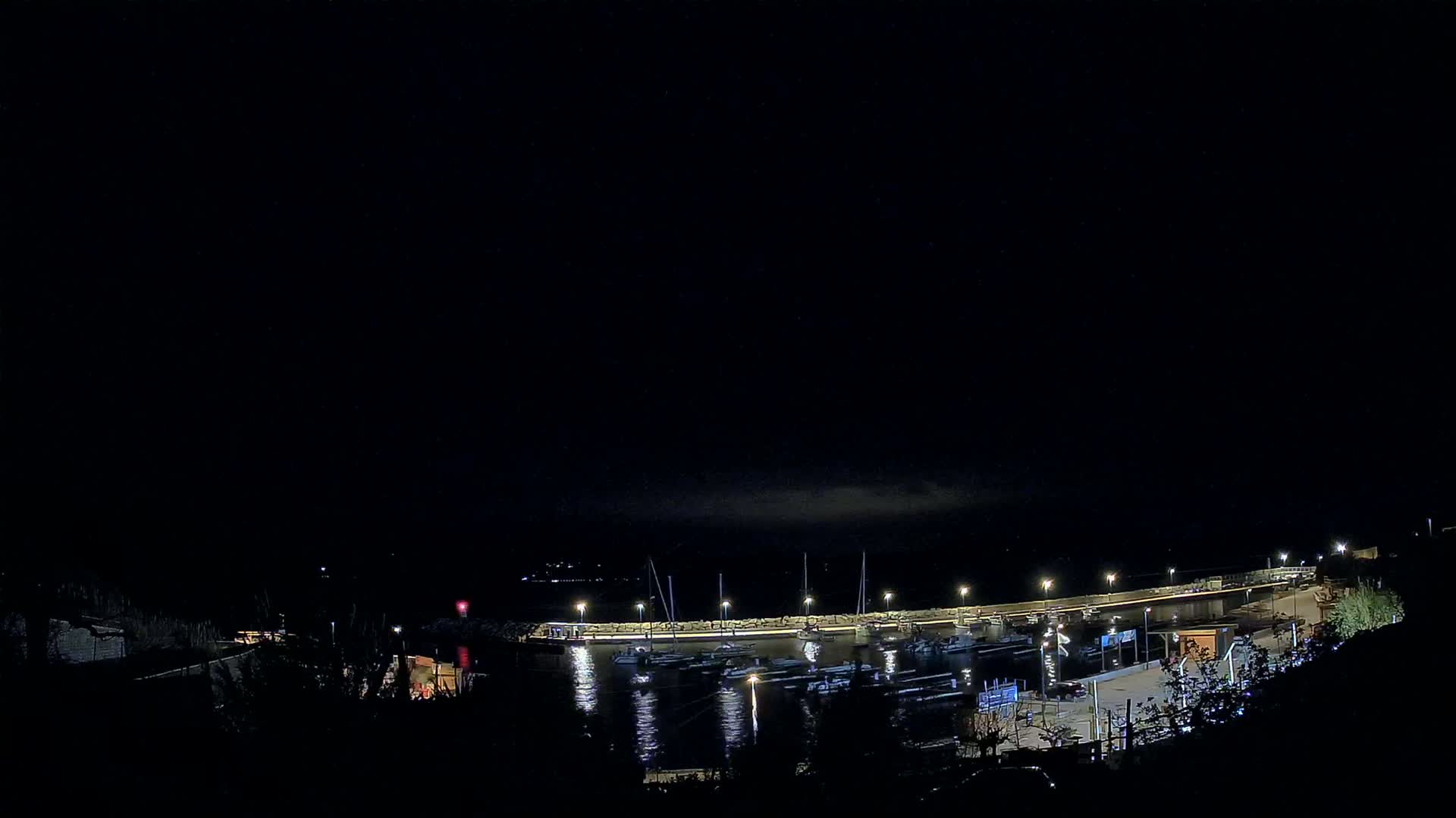 A nighttime view of a harbor with boats moored at a lit dock under a dark, mostly clear sky.