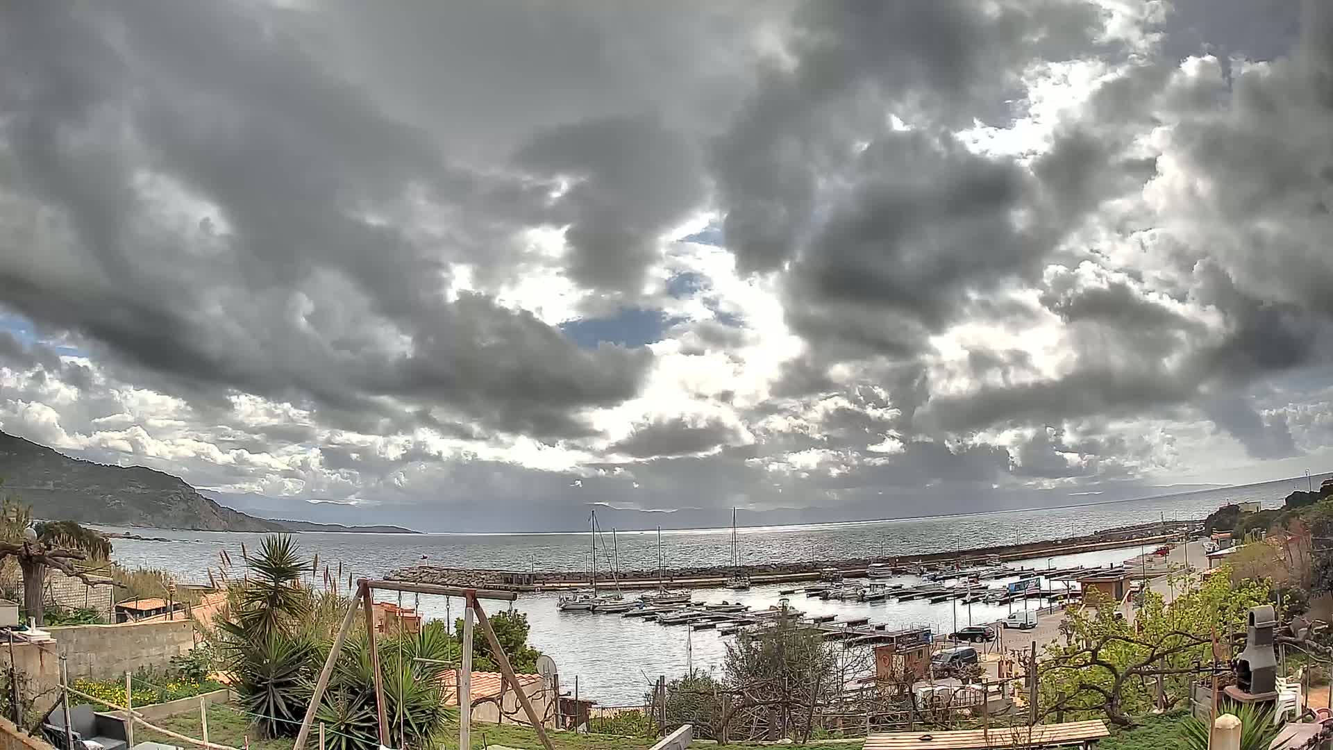 A small harbor filled with boats is visible under a mostly cloudy sky, with hints of sunlight breaking through the clouds, and a hilly coastline in the background.