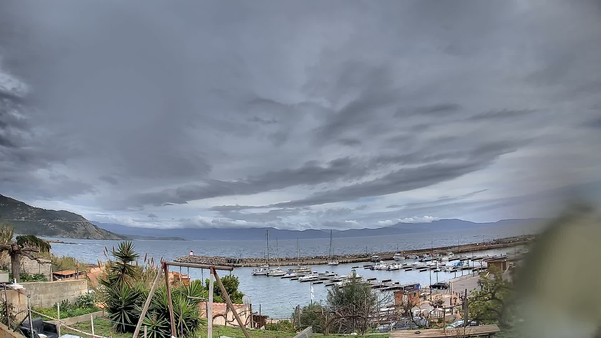 A small harbor filled with boats is nestled between a rocky coastline and a calm sea under a heavily clouded sky.