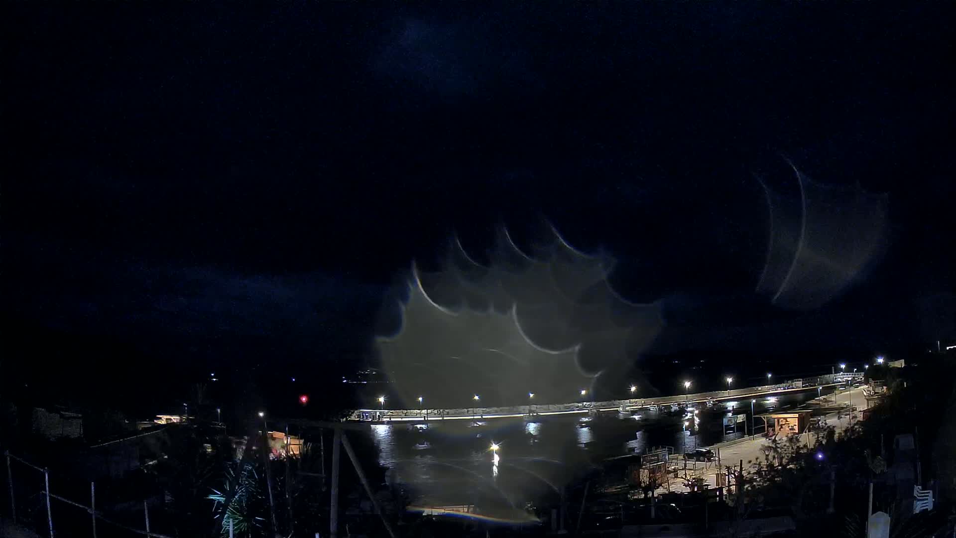 A nighttime view of a harbor with boats, illuminated by lights, under a dark, cloudy sky.