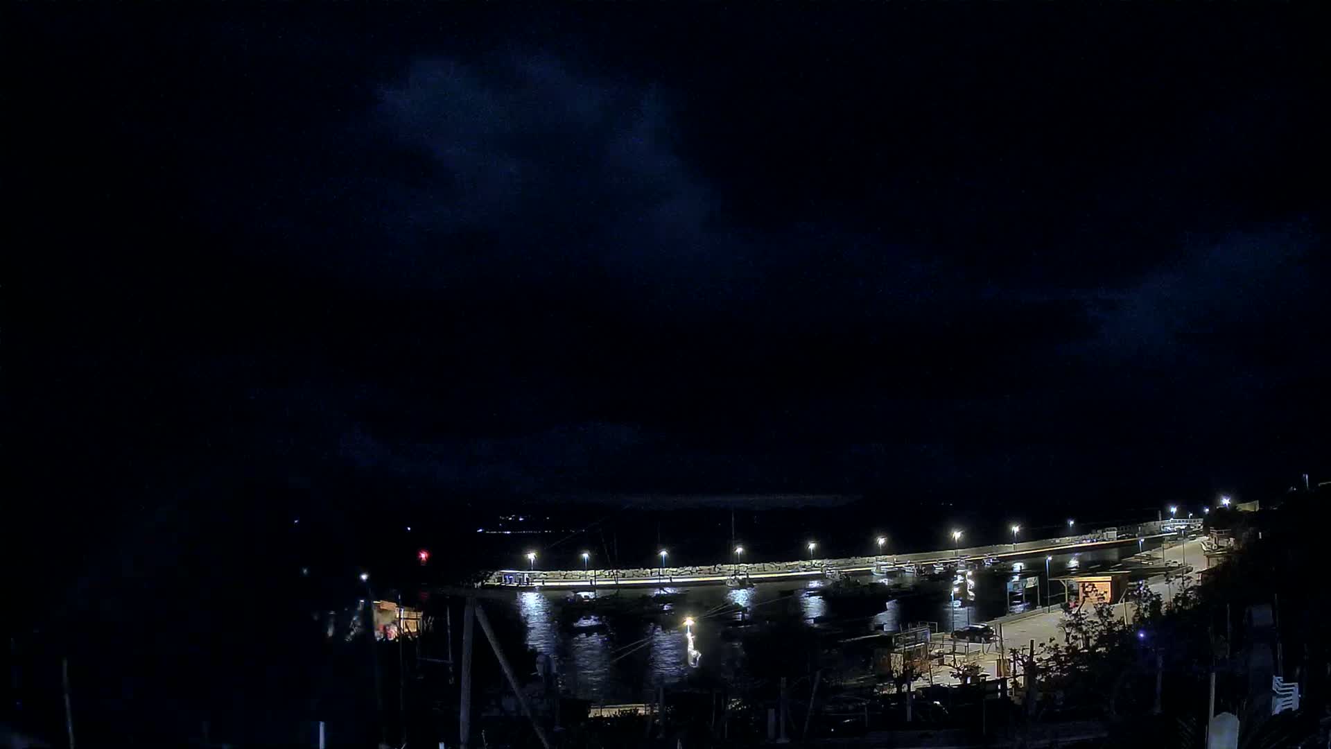 A nighttime view of a harbor with boats moored at a lit pier under a dark, cloudy sky.