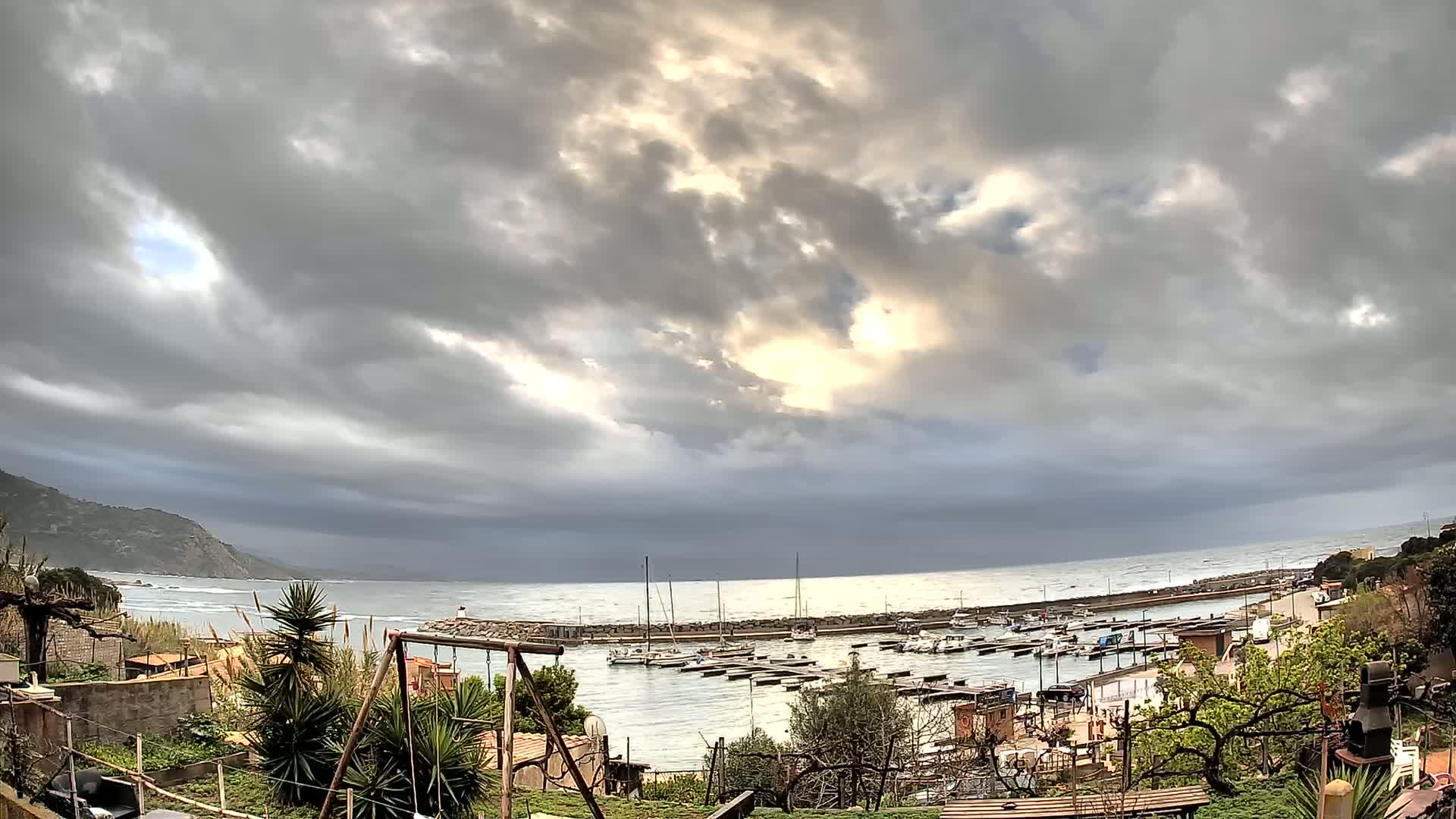 A small harbor filled with boats is seen from a hillside overlooking a partly cloudy, overcast ocean with dark, heavy clouds.