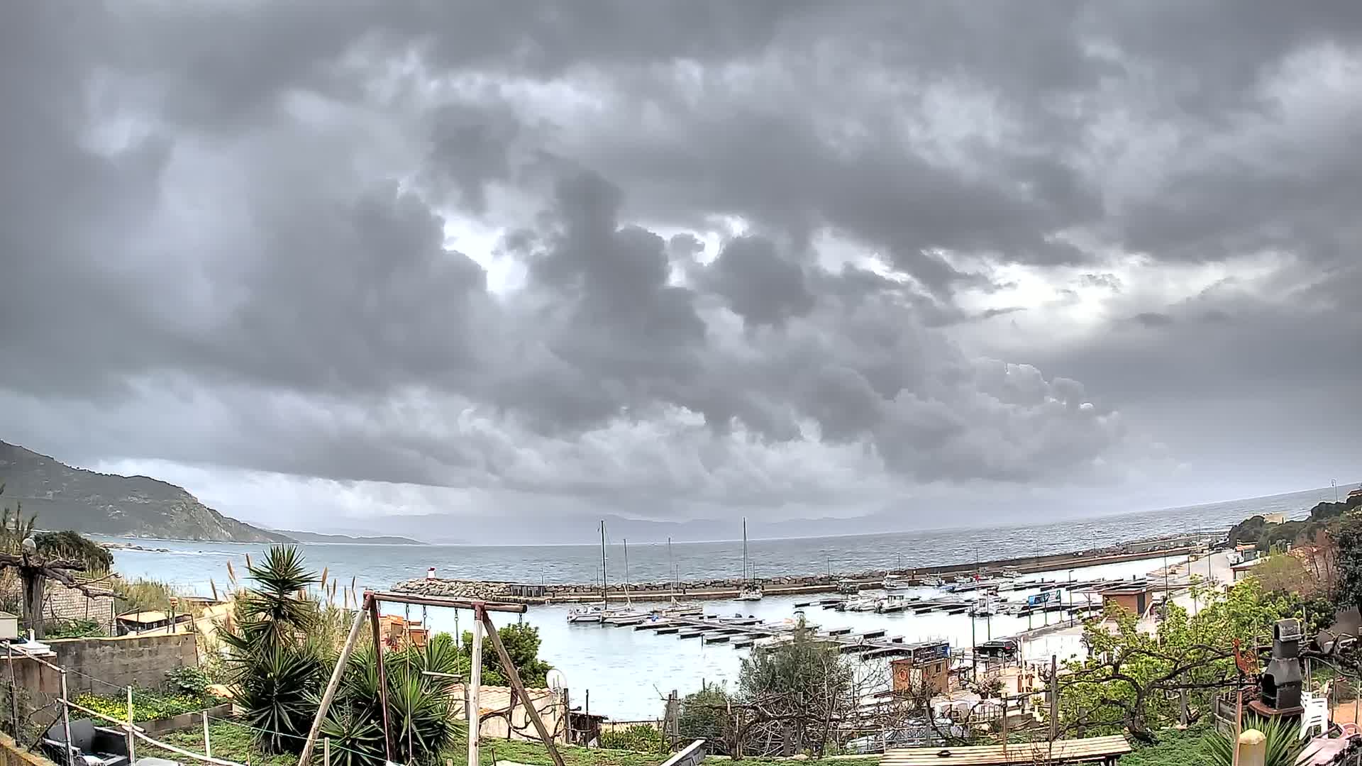 A small marina filled with boats is situated on a calm sea next to a rocky coastline under a heavily overcast sky.