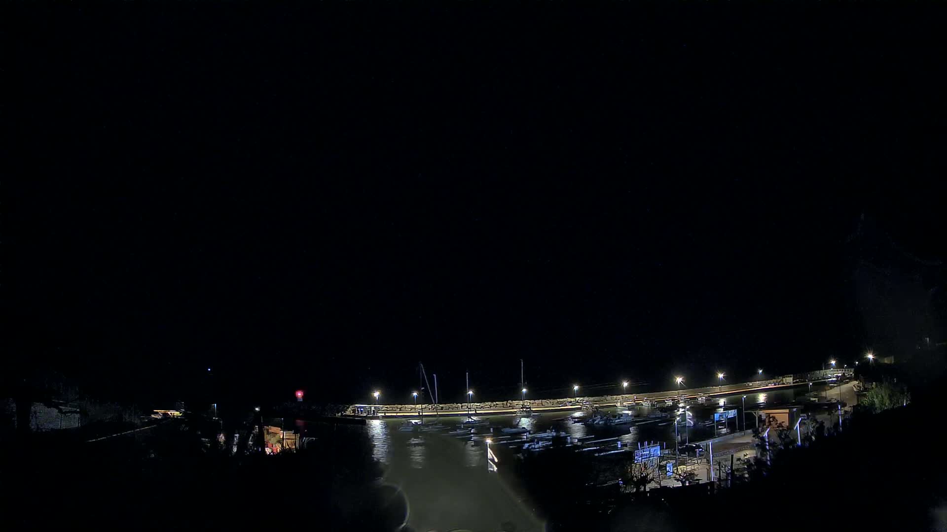 A nighttime view of a harbor with several boats docked, illuminated by lights along a seawall under a dark sky.