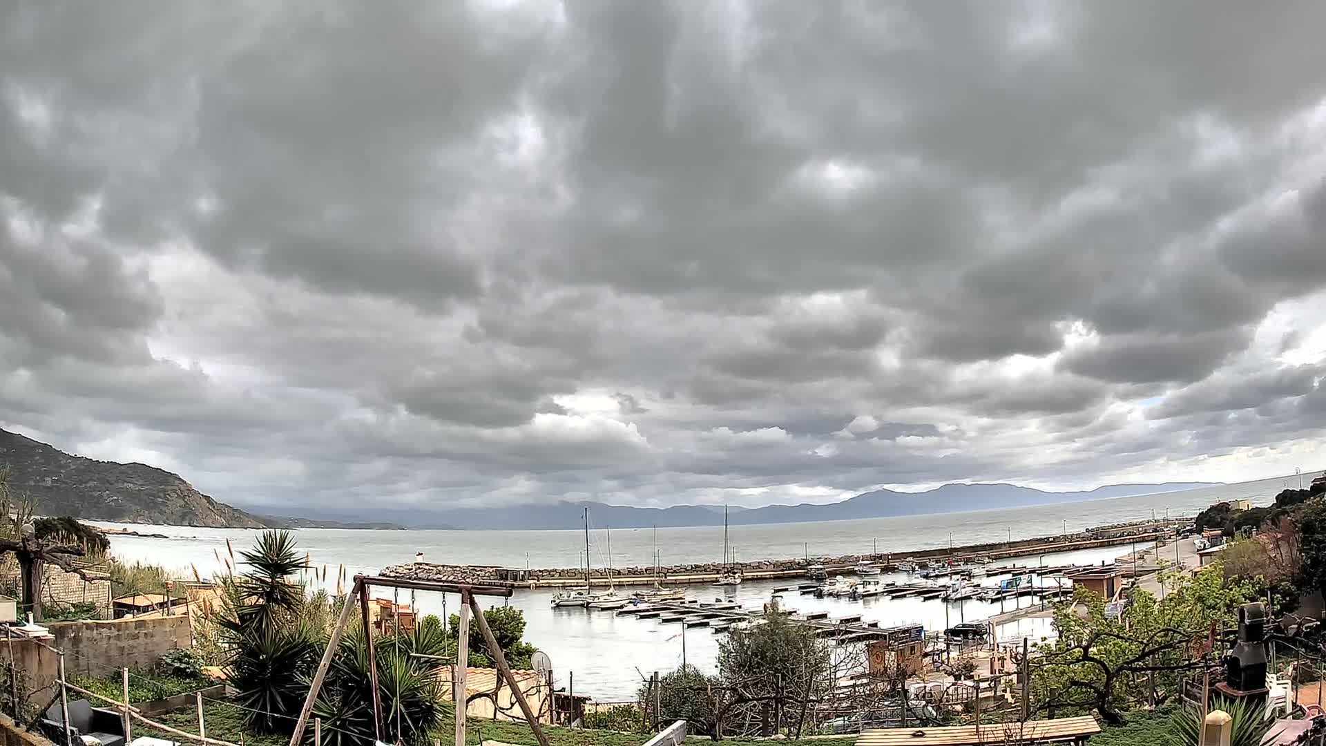 A small harbor filled with boats is seen from a hillside under a heavily clouded sky.