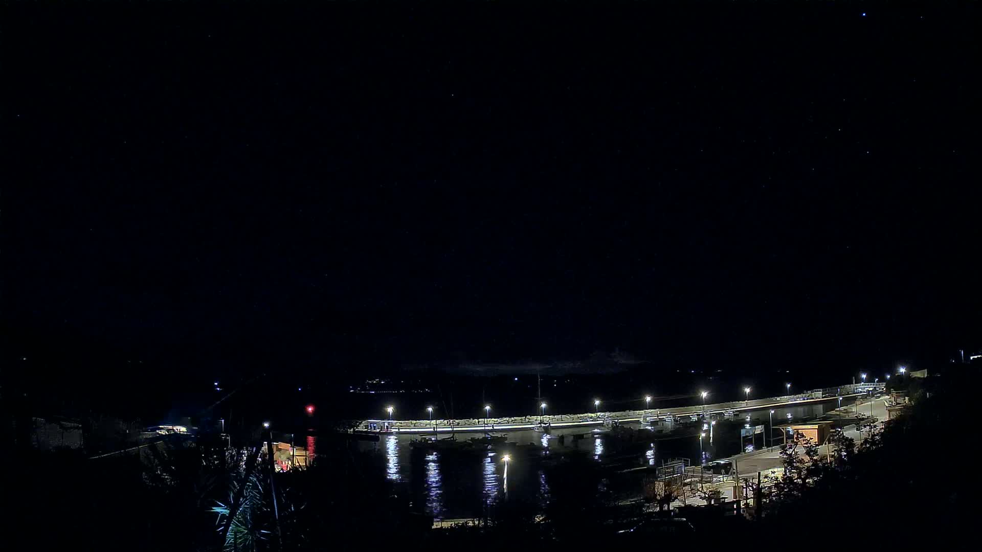 A nighttime view of a harbor with boats docked along a lit pier under a clear, starry sky.