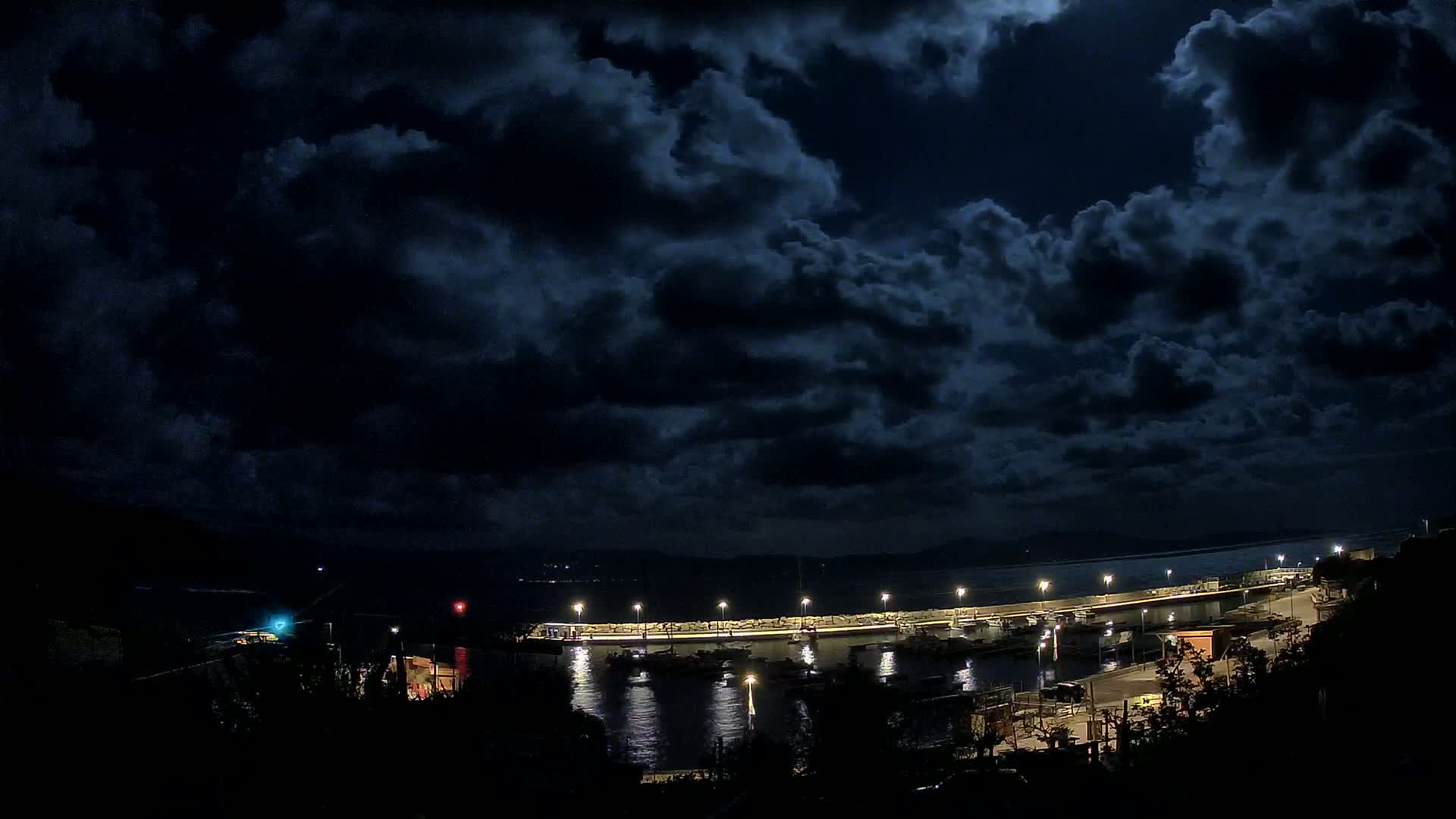 A nighttime harbor scene under a dark, cloudy sky, showing a lit breakwater with boats moored alongside it.