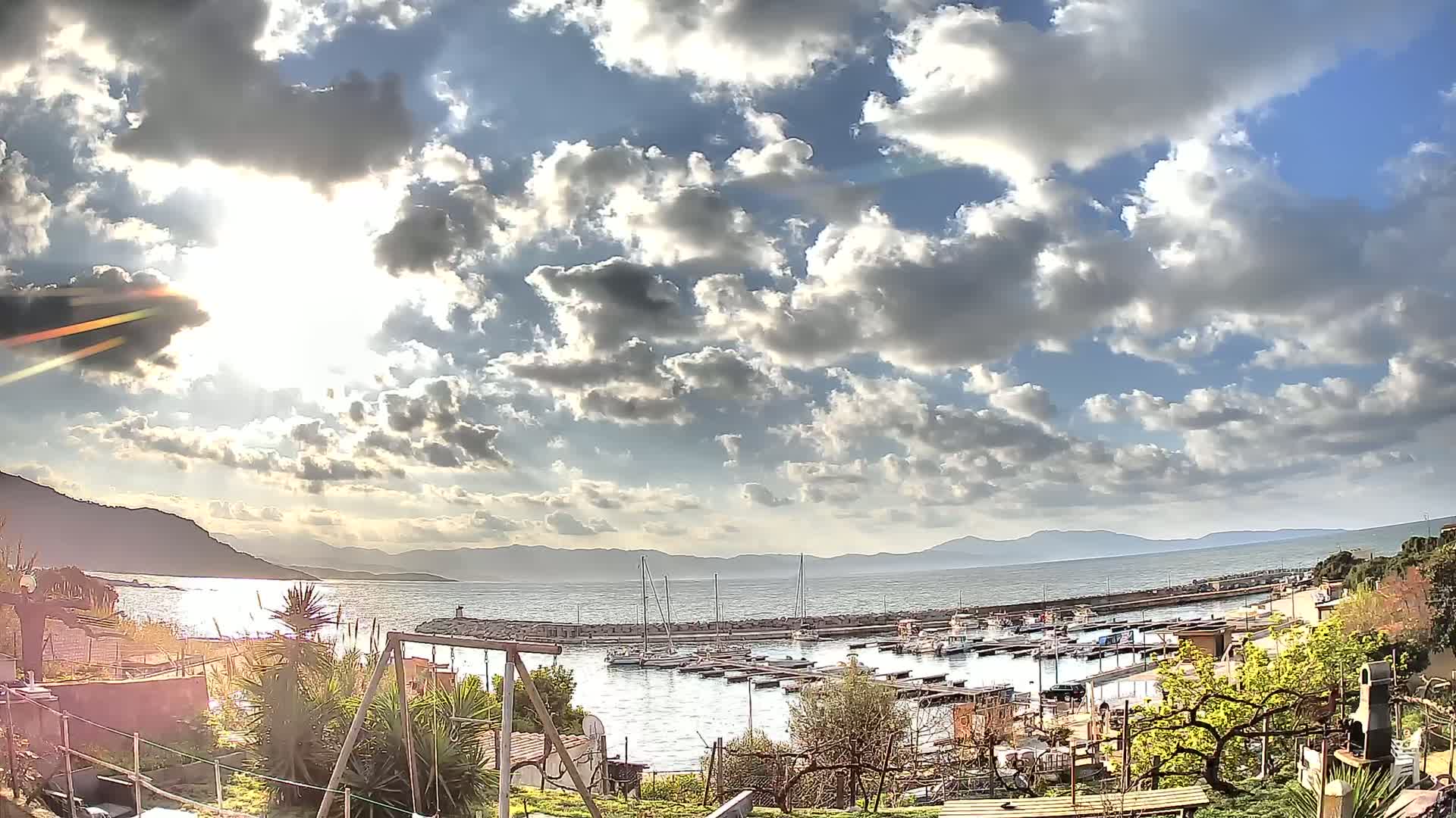 A partly sunny day reveals a small marina filled with boats, nestled between hills and a calm body of water under a partly cloudy sky.