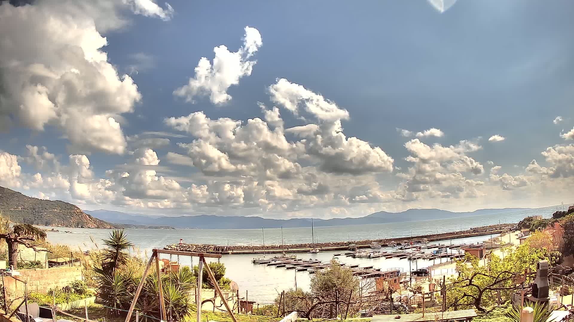 A small marina filled with boats sits on a calm bay under a partly cloudy sky, with hills visible in the distance.