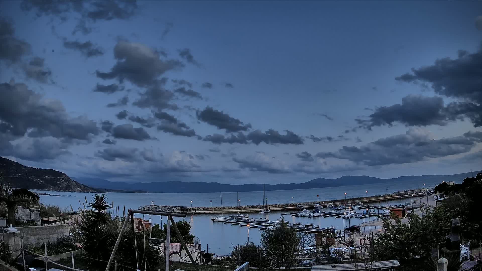 A small marina filled with boats is situated on a calm sea, under a mostly cloudy twilight sky, with mountains visible in the distance.