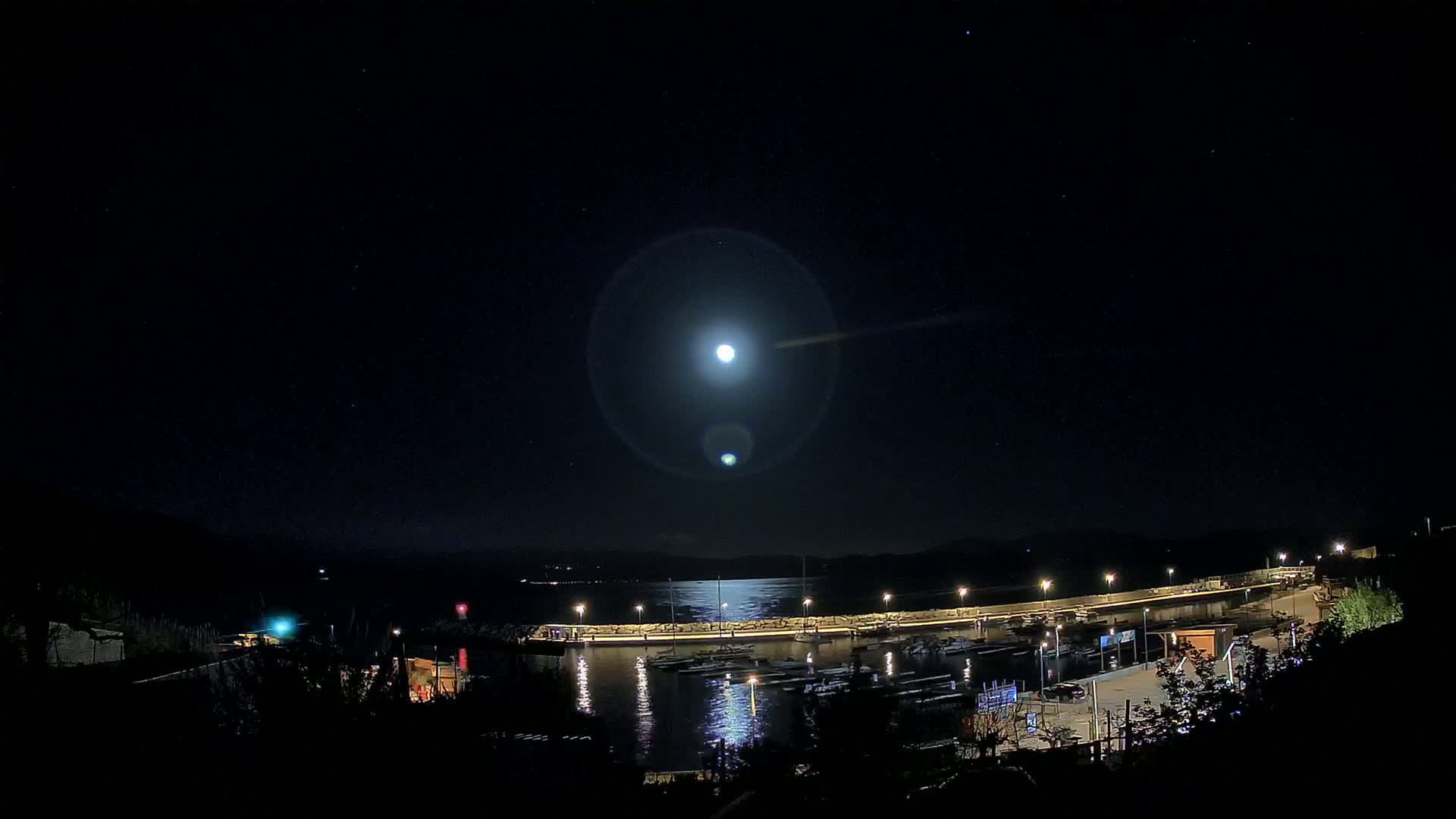 A bright moon illuminates a harbor filled with boats at night, under a clear, starry sky.